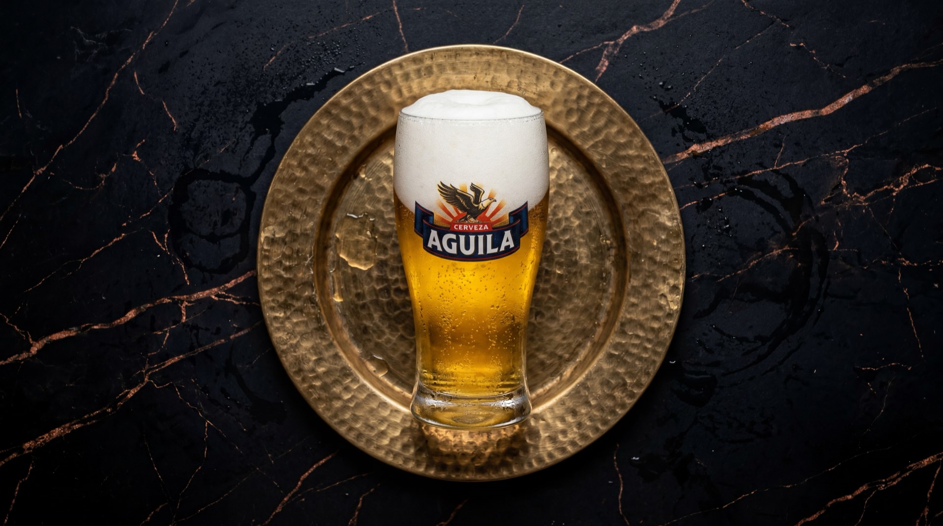 Cerveza Aguila de Bavaria, botella dorada con etiqueta del aguila sobre mesa de marmol oscuro con vetas doradas