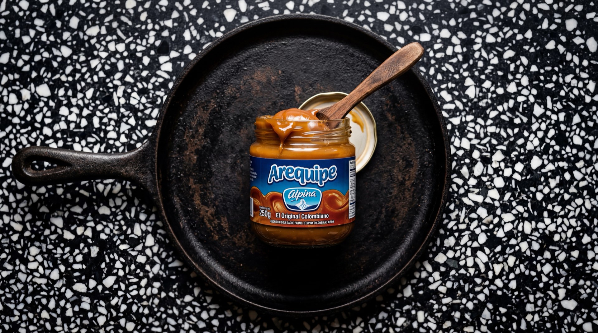 Arequipe Alpina en tarro de vidrio abierto con cucharada de dulce de leche colombiano sobre mesa de marmol oscuro