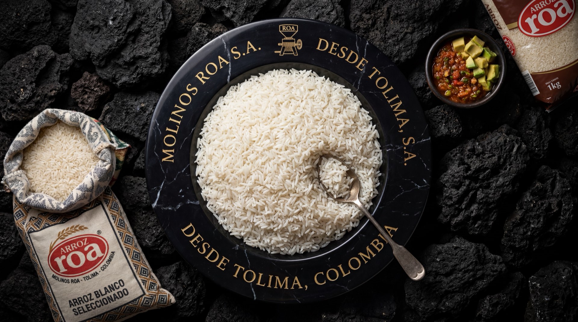 Paquete de arroz Roa blanco junto a taza de arroz cocido suelto sobre superficie de marmol oscuro