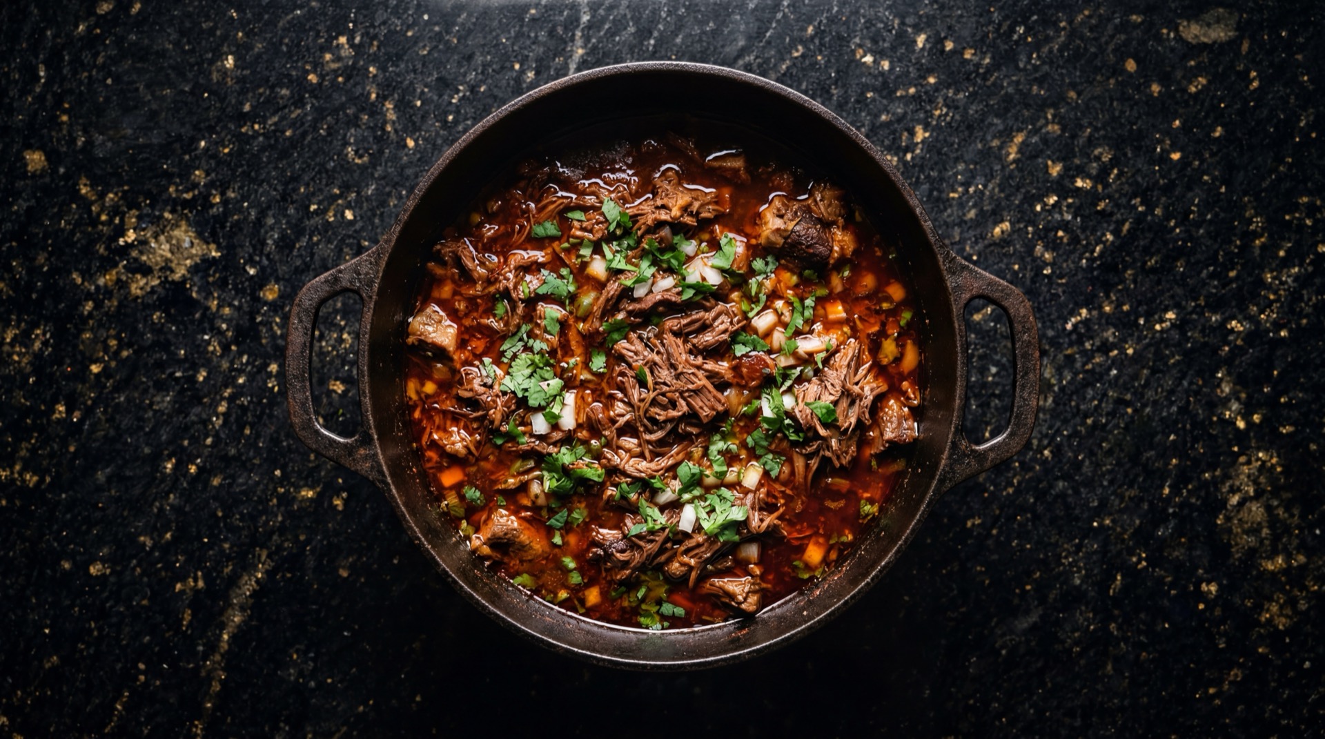 Birria de res de Jalisco en plato hondo con consome rojo intenso, carne deshebrada y cilantro fresco picado sobre mesa de granito negro