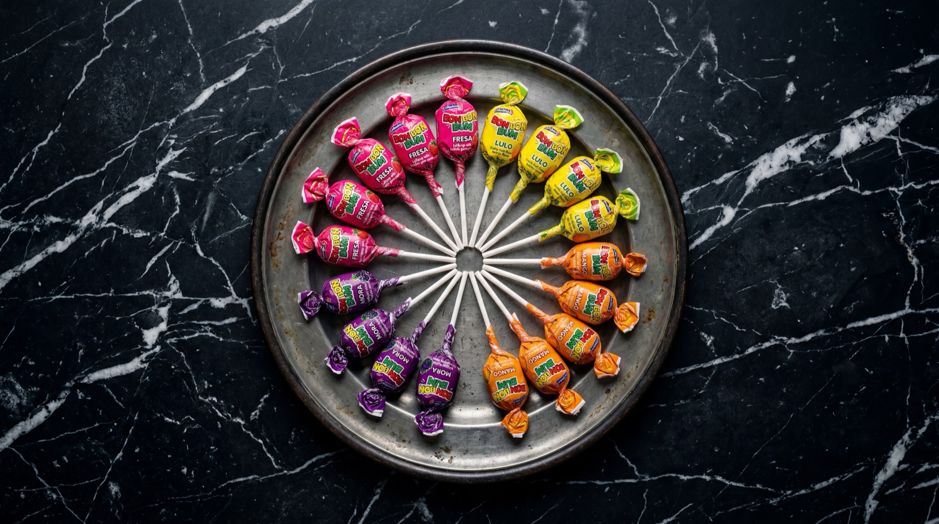 Bon Bon Bum cortado a la mitad mostrando la capa exterior de caramelo duro de fresa rosado y el centro de chicle blanco, junto a chupetes enteros de varios sabores de Colombina