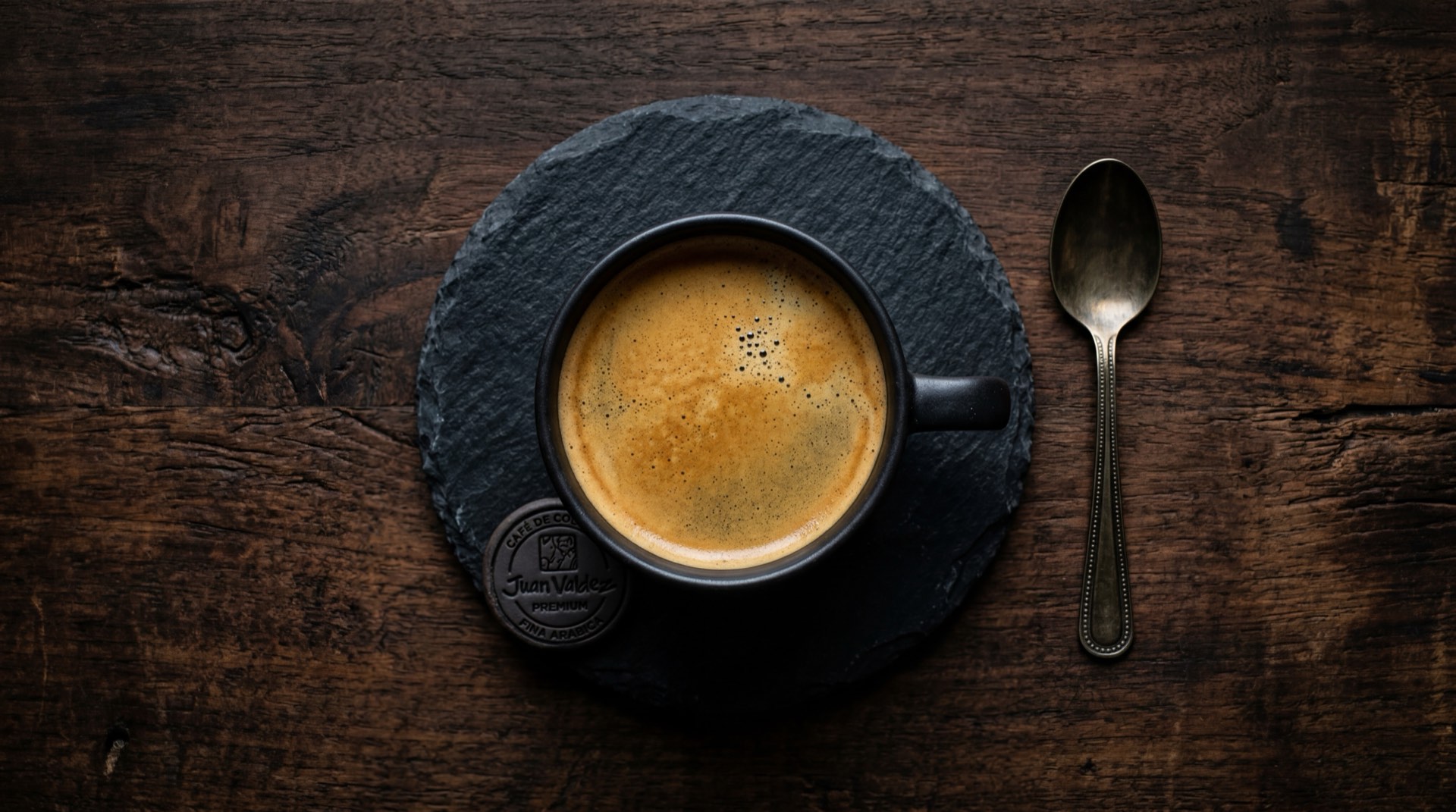Taza de cafe negro Juan Valdez recien preparado sobre plato de ceramica oscura en mesa de marmol negro con vetas doradas