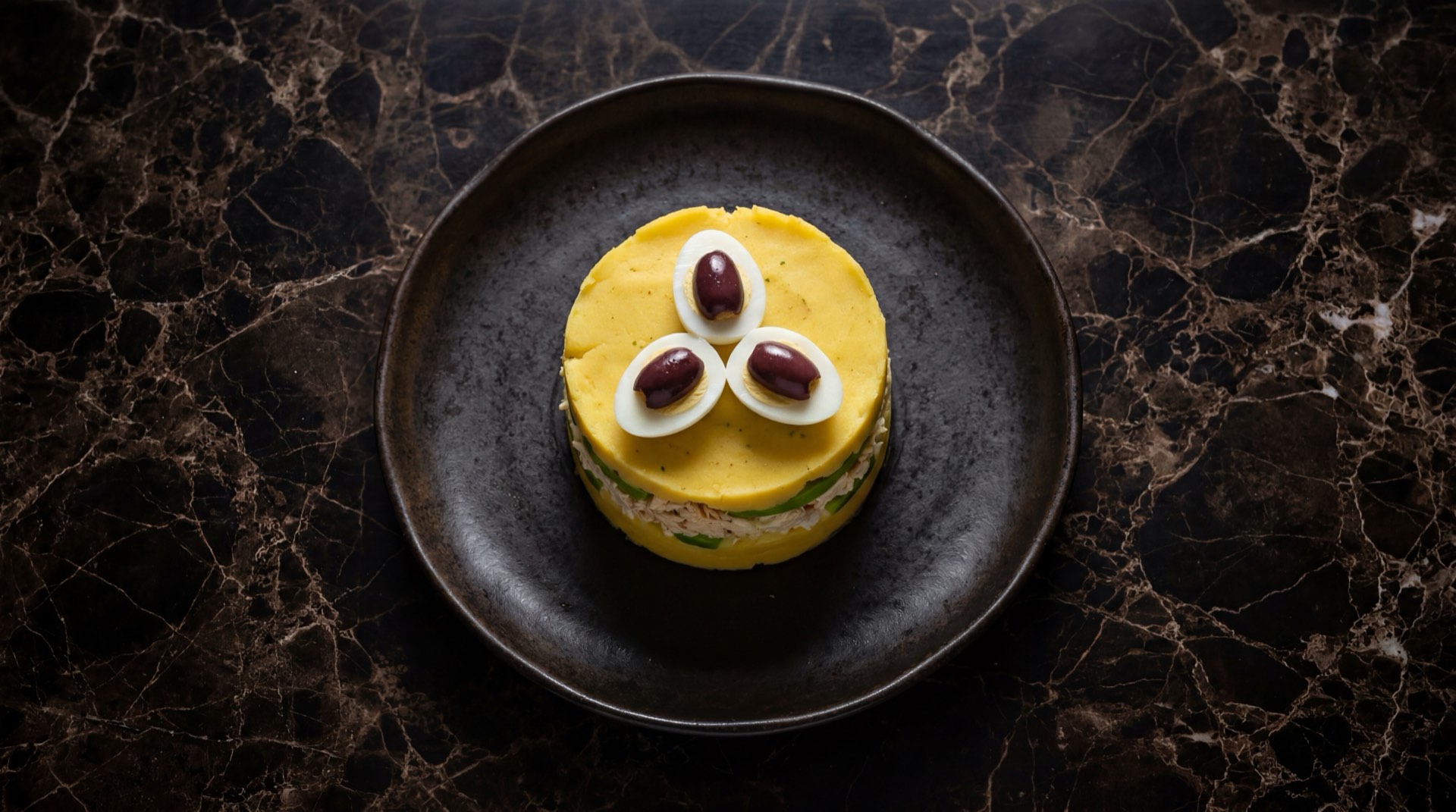 Causa limena peruana clasica de pollo con capas de papa amarilla prensada, palta Hass, pollo deshilachado con mayonesa, huevo duro y aceitunas de botija negras en plato de vidrio ahumado sobre mesa de cuarcita negra