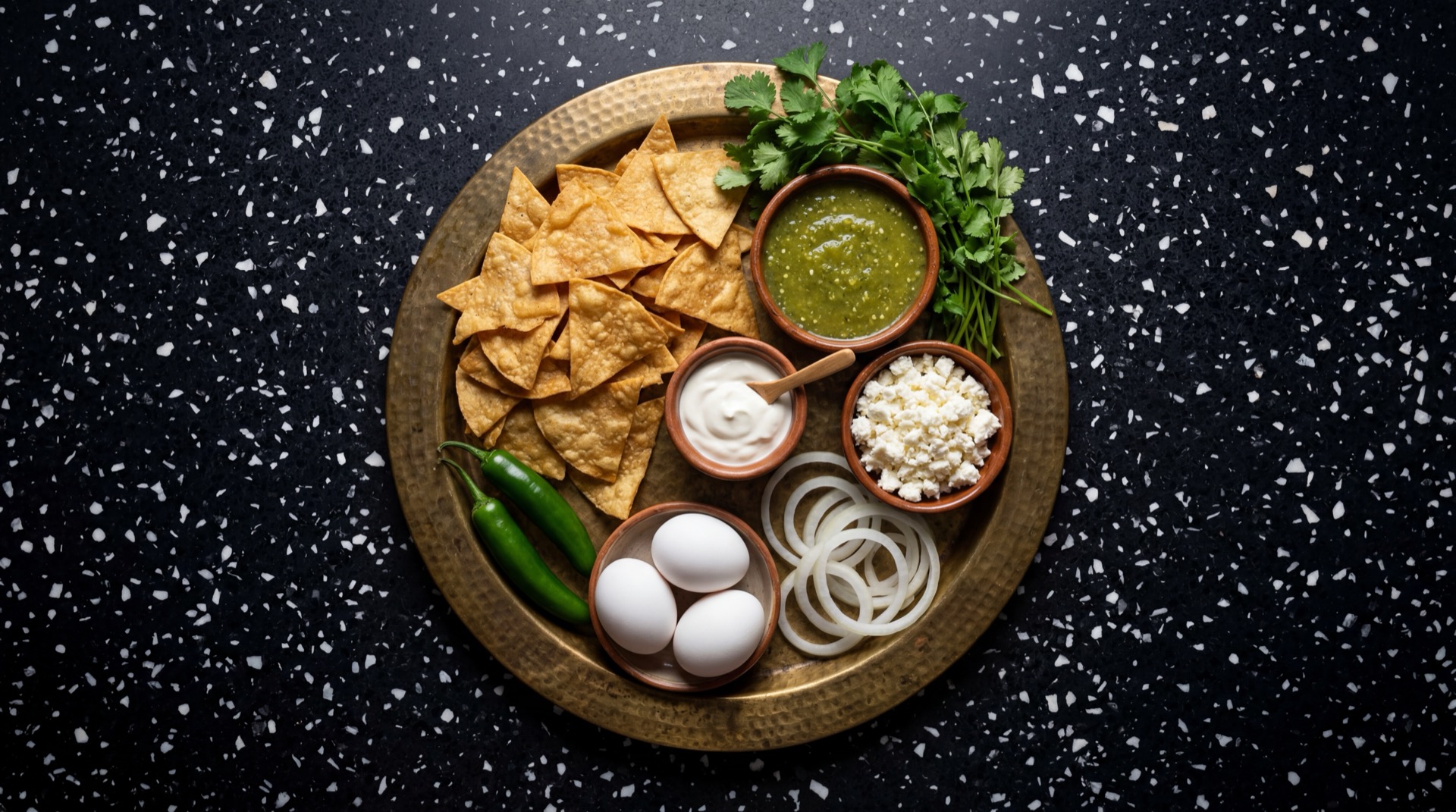 Ingredientes de los chilaquiles mexicanos: totopos de tortilla de maiz fritos, tomatillos verdes frescos sin cascara, chiles serranos frescos verdes, crema agria mexicana, queso fresco desmoronado, cebolla blanca en rodajas, cilantro fresco picado, dientes de ajo pelados y sal de mesa