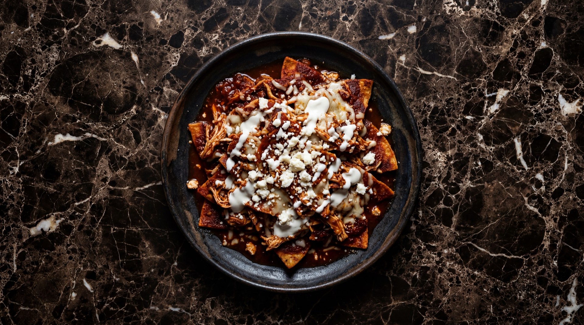 Chilaquiles rojos mexicanos con huevo frito estrellado, crema acida, queso fresco desmenuzado y cebolla blanca en rodajas sobre plato de hierro oscuro en mesa de basalto volcanico