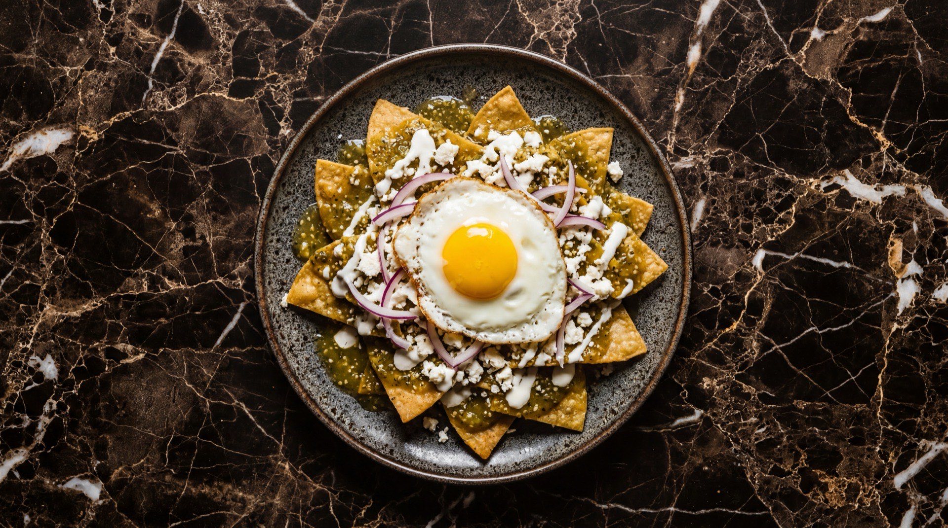 Chilaquiles verdes mexicanos con totopos de tortilla de maiz fritos banados en salsa verde de tomatillo, crema agria mexicana, queso fresco desmoronado y cilantro fresco picado en plato de ceramica negra mate sobre mesa de marmol negro con vetas doradas
