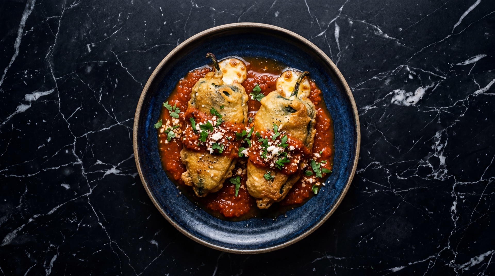 Chiles rellenos de queso Oaxaca capeados en huevo dorado con salsa de tomate rojo en plato de ceramica oscura sobre mesa de marmol negro con vetas doradas
