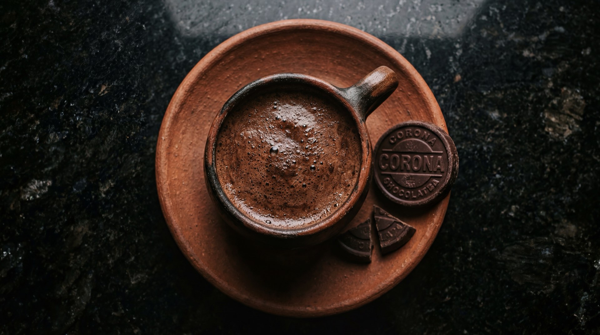 Pastillas de chocolate Corona colombiano sobre superficie oscura junto a taza de chocolate caliente