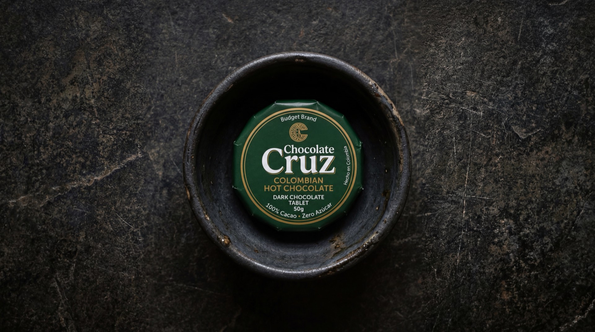 Pastilla de Chocolate Cruz de Nacional de Chocolates, empaque verde sobre mesa de marmol oscuro con taza de chocolate caliente