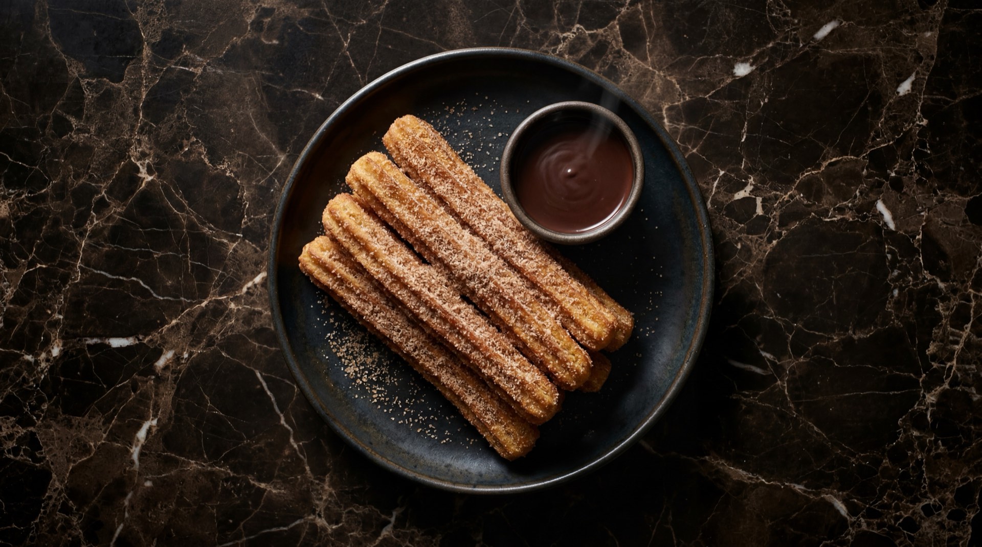 Churros mexicanos dorados cubiertos de azucar con canela molida en plato de hierro oscuro sobre mesa de marmol emperador