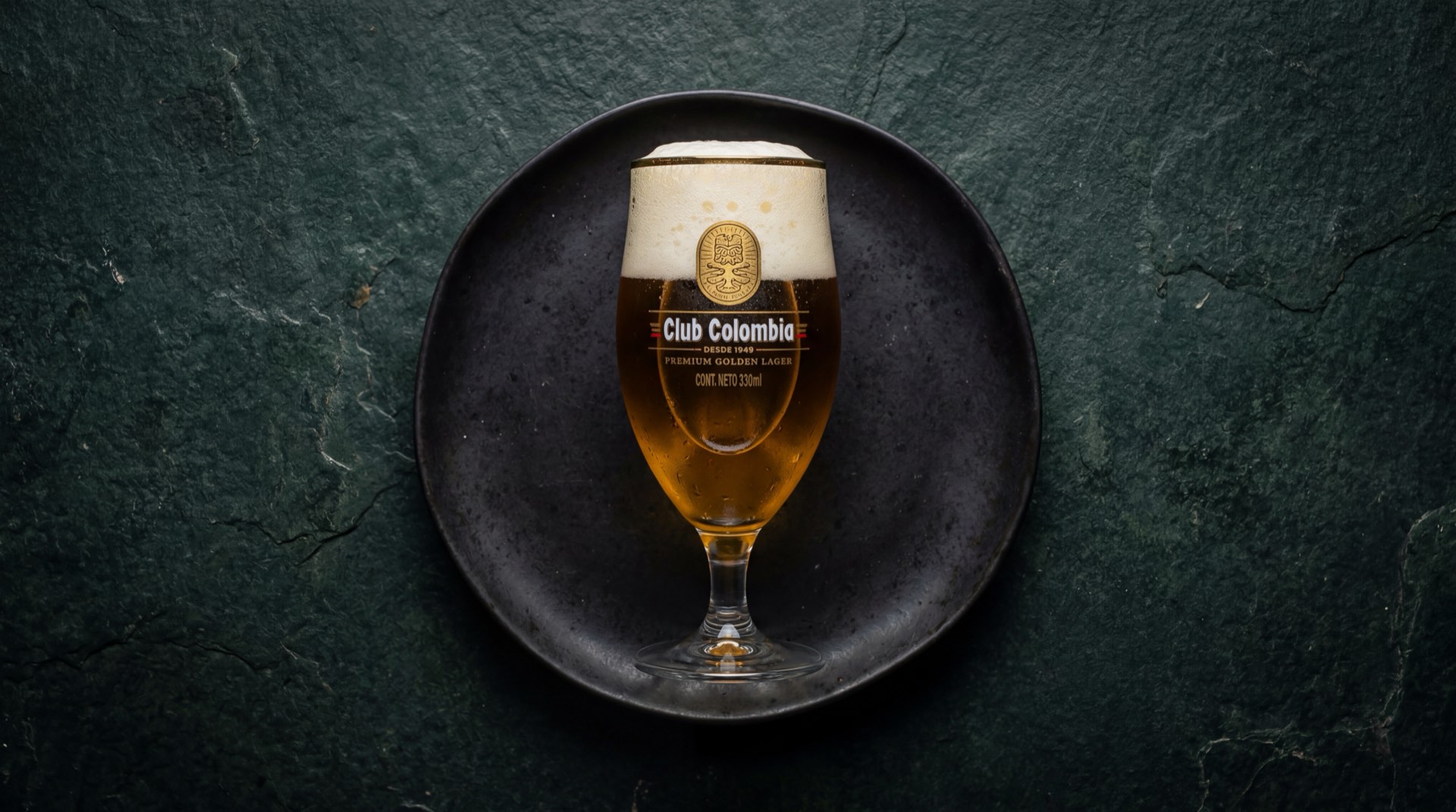 Botella de cerveza Club Colombia Dorada con etiqueta dorada sobre mesa de marmol negro, fotografia oscura de producto premium
