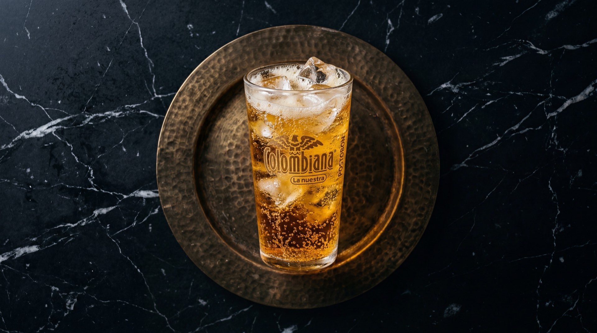 Colombiana Postobon botella de vidrio con liquido dorado ambar gasificado sobre mesa de granito negro con destellos dorados