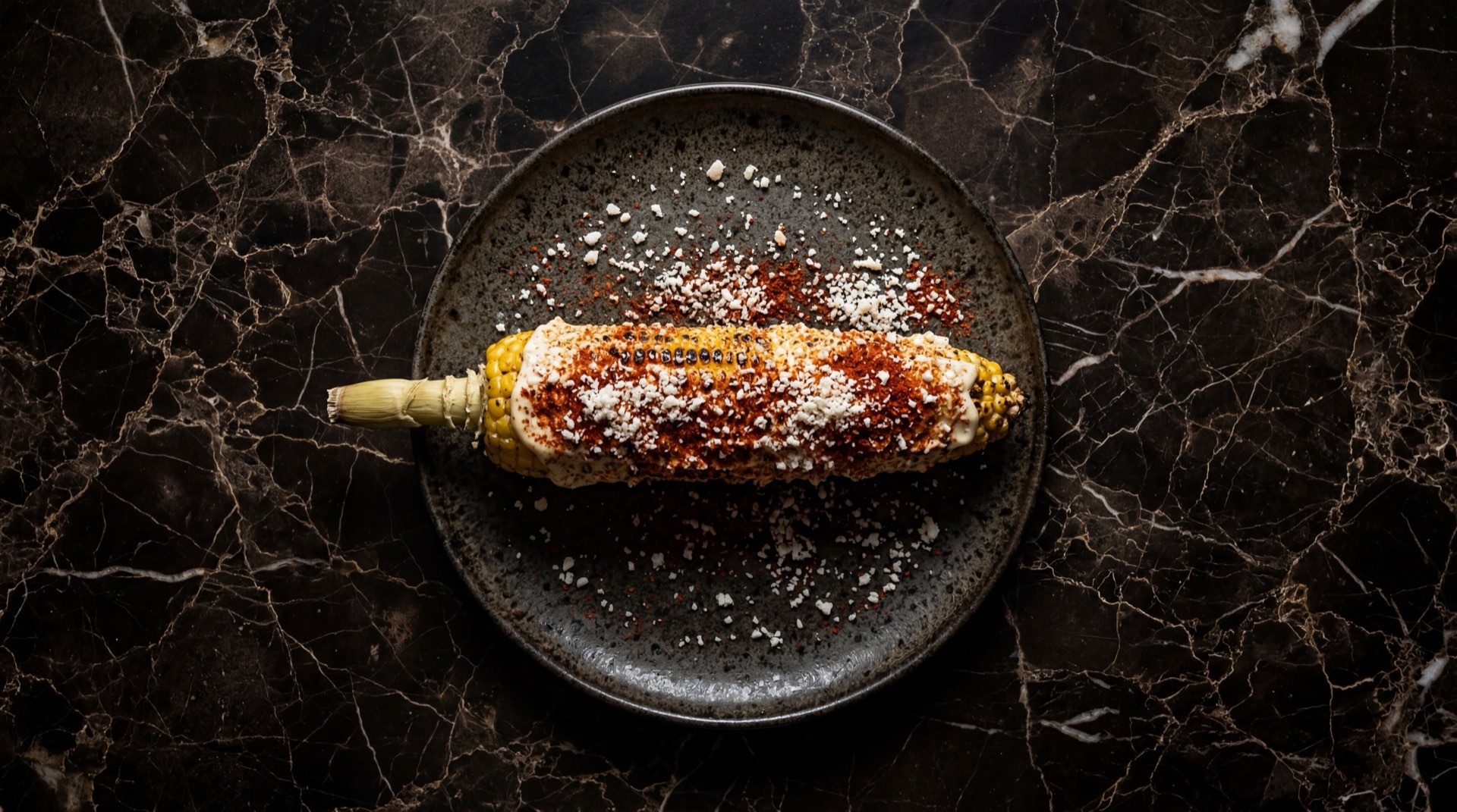 Elote mexicano preparado con mayonesa, chile en polvo rojo, queso cotija rallado y limon verde sobre plancha de hierro fundido