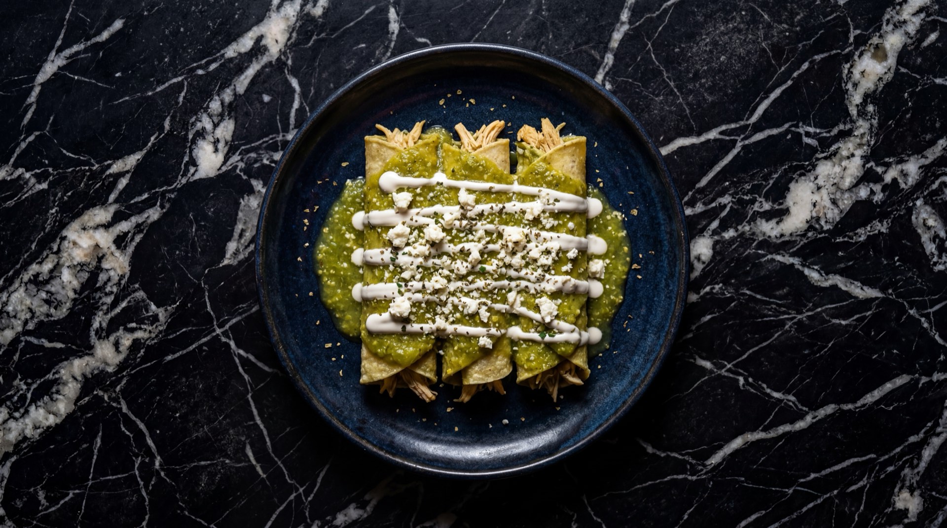 Enchiladas verdes mexicanas de pollo banadas en salsa de tomatillo con crema agria y queso fresco desmoronado sobre plato de ceramica oscura