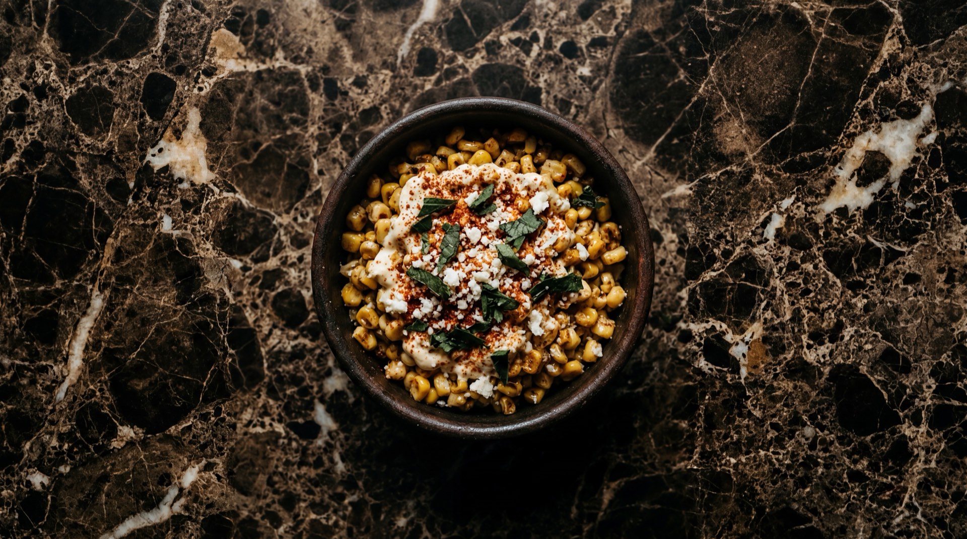 Esquites mexicanos en vaso con mayonesa, chile en polvo rojo, queso cotija rallado y limon verde sobre superficie de granito negro
