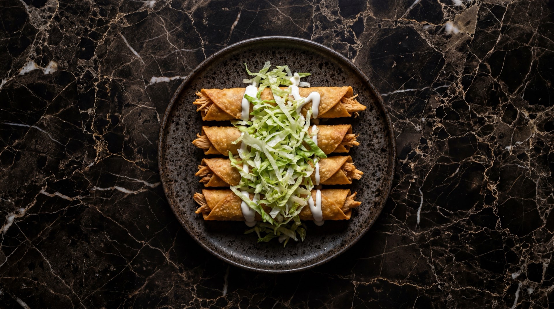 Flautas mexicanas de pollo deshebrado doradas y crujientes con crema acida y lechuga sobre plato de obsidiana negra en mesa de marmol negro con vetas blancas