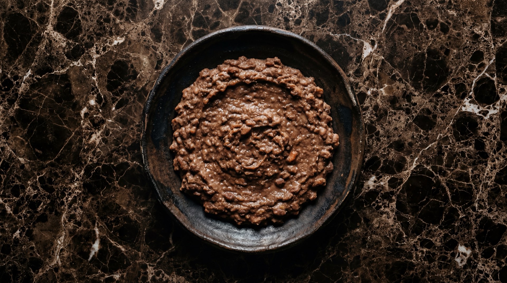 Frijoles refritos mexicanos con queso fresco desmoronado en plato de barro sobre mesa de madera oscura