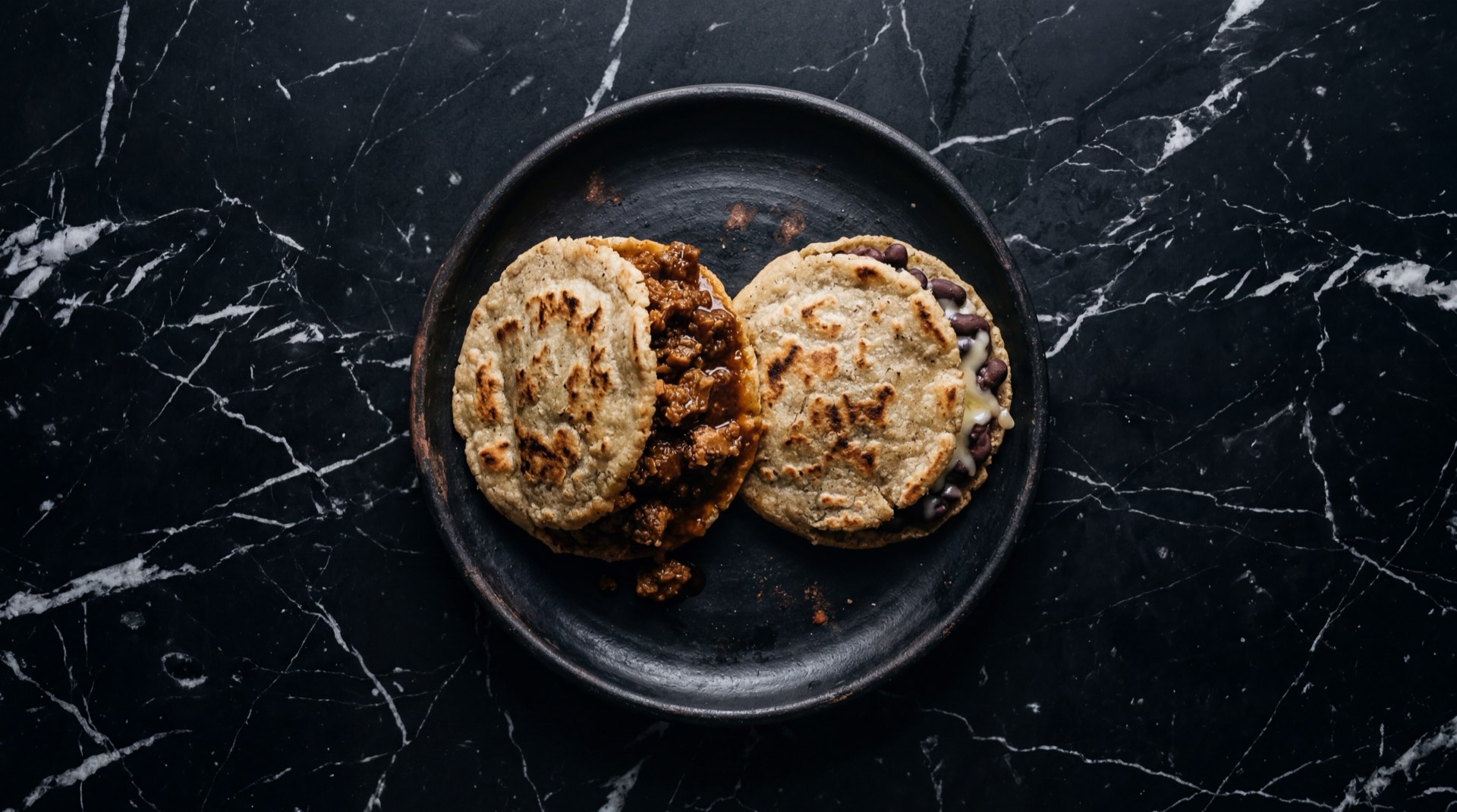Gorditas mexicanas doradas rellenas de chicharron prensado sobre plato de barro oscuro en mesa de marmol negro con vetas doradas