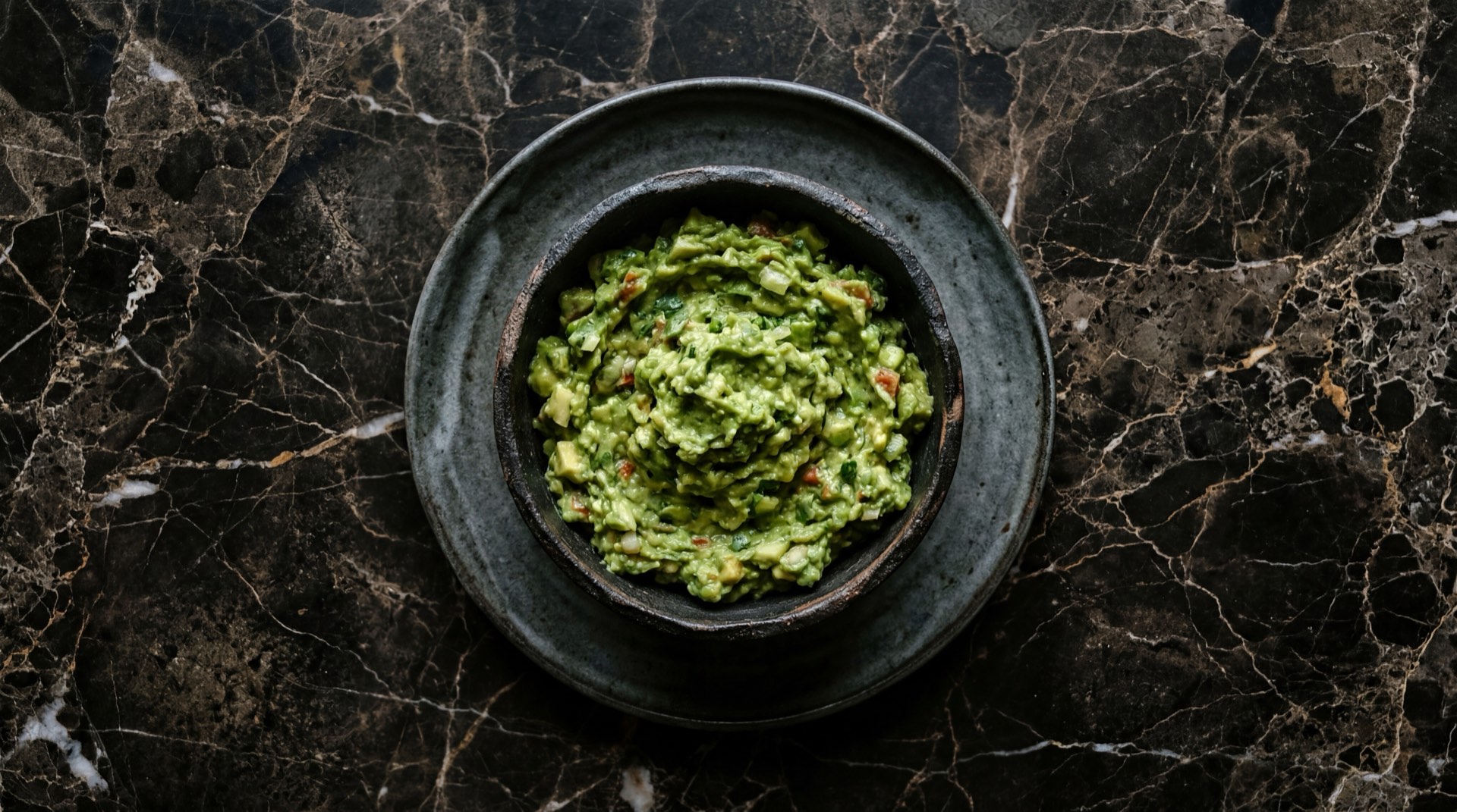 Guacamole mexicano fresco con totopos de maiz en molcajete de piedra volcanica sobre mesa de granito negro