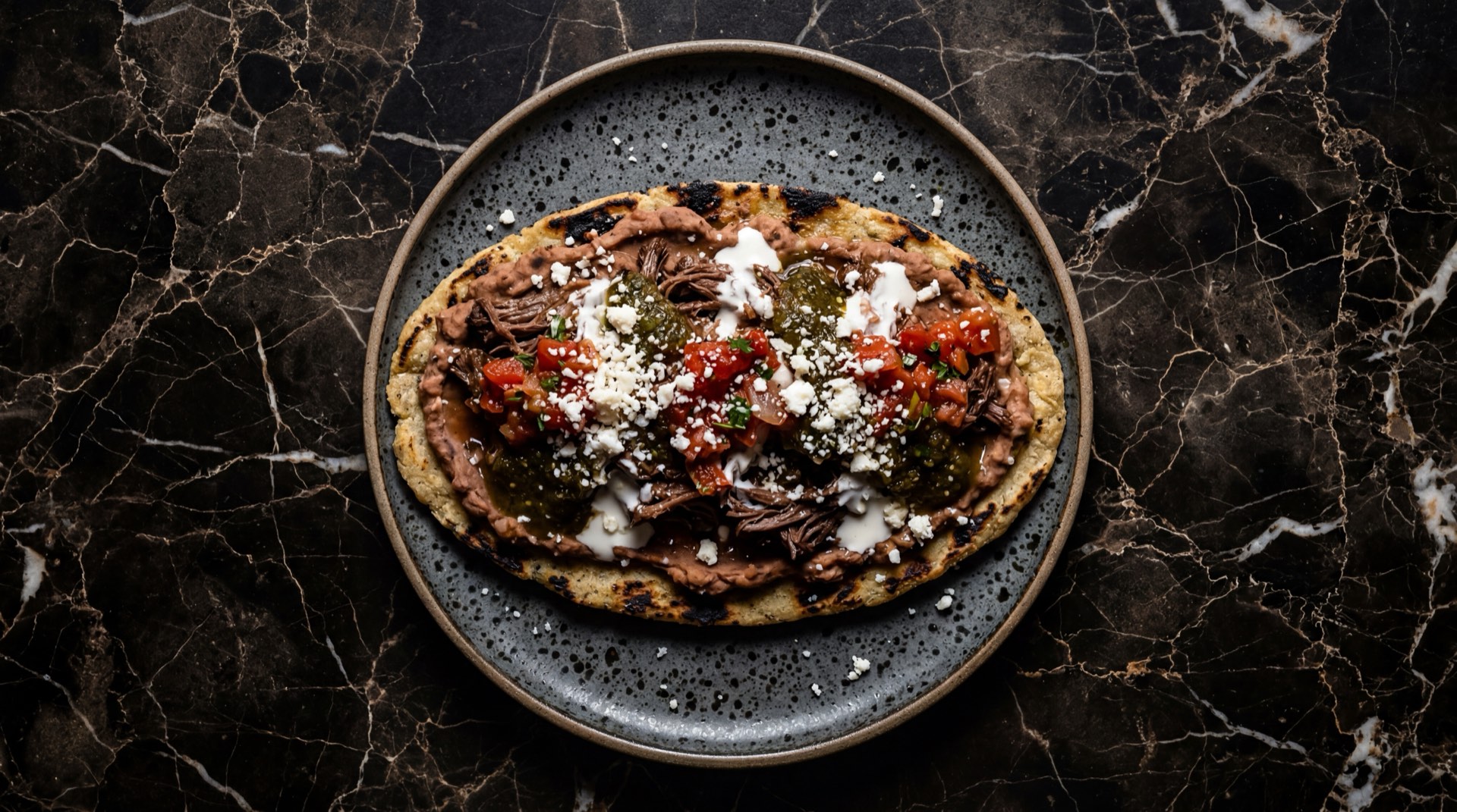 Huaraches mexicanos con bistec de res sobre frijoles refritos negros en plato de barro negro sobre mesa de granito negro con vetas doradas
