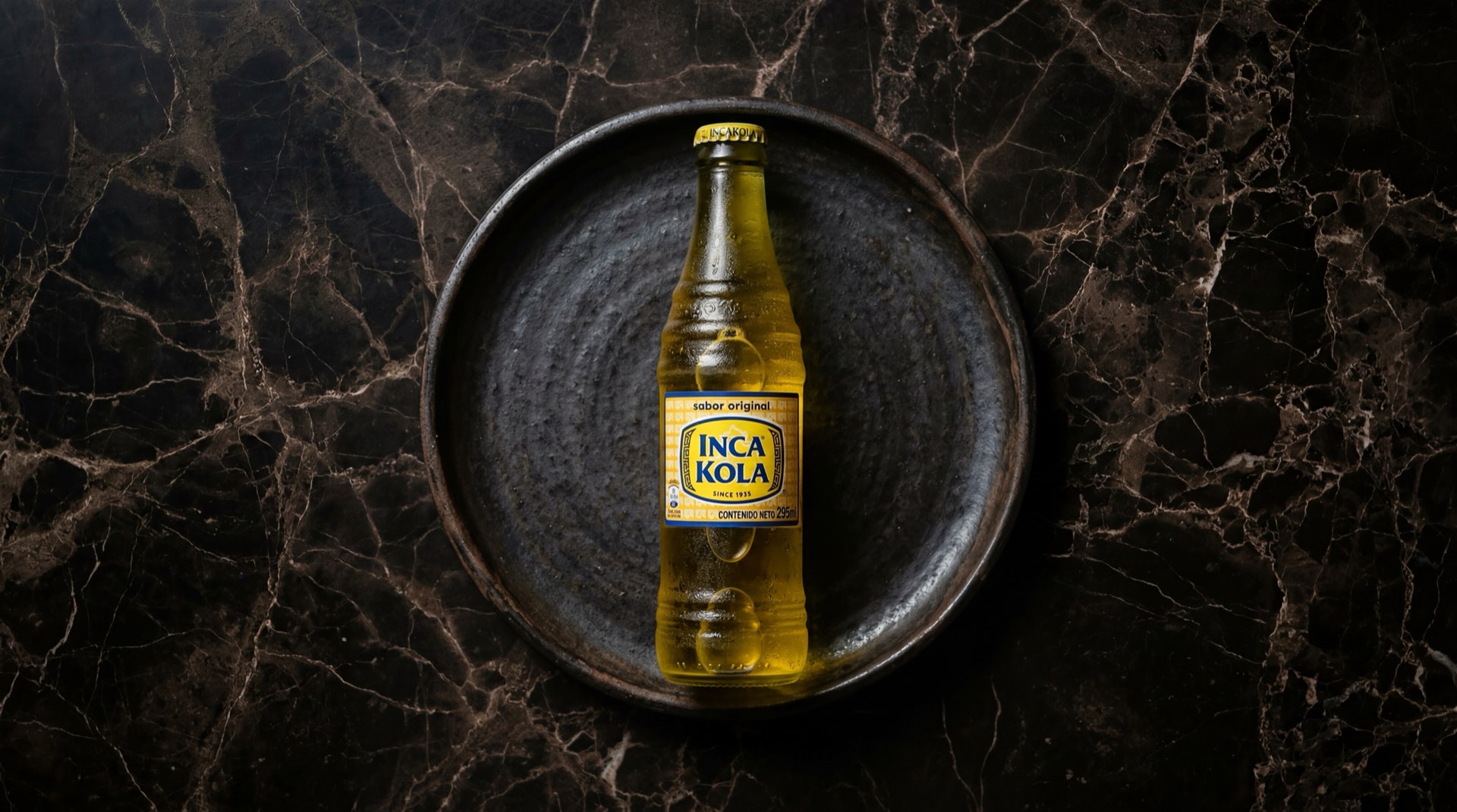 Botella de Inca Kola peruana dorada con vaso de cristal lleno de gaseosa dorada con hielo sobre plato de acero ennegrecido en mesa de marmol azul medianoche