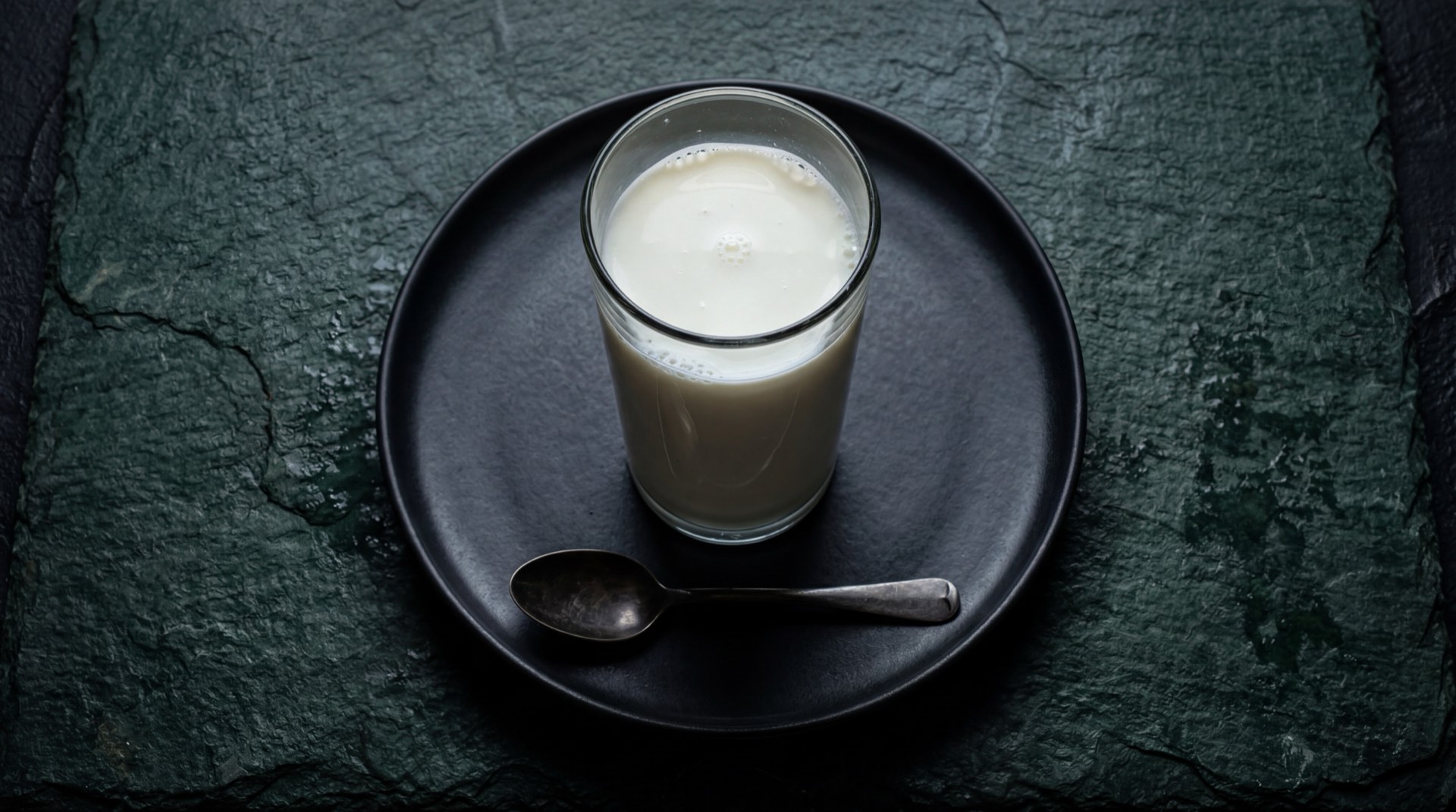 Vaso de leche Alqueria entera fresca sobre mesa de marmol negro con empaque azul y blanco de la marca