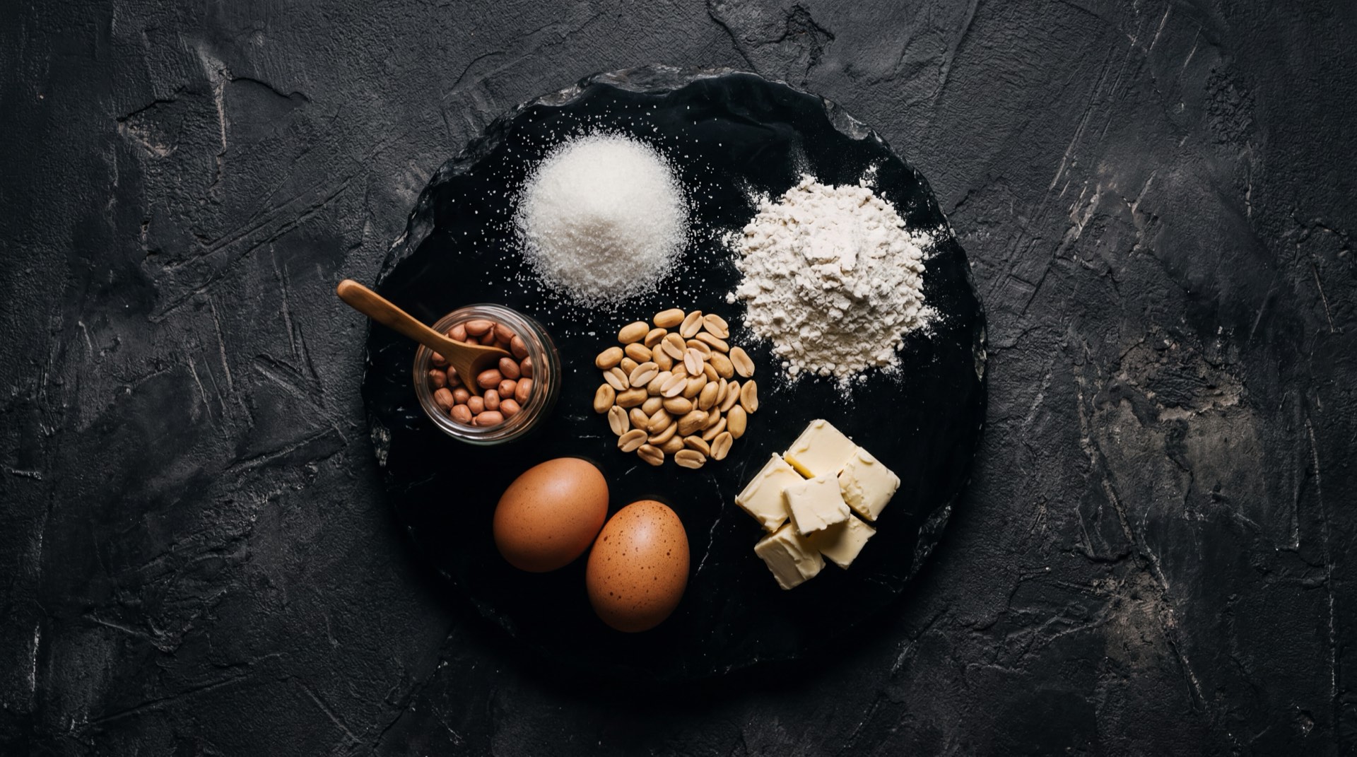 Ingredientes de la galleta Mana de Ramo: mani tostado entero, harina de trigo refinada, azucar blanca granulada y mantequilla vegetal sobre superficie oscura