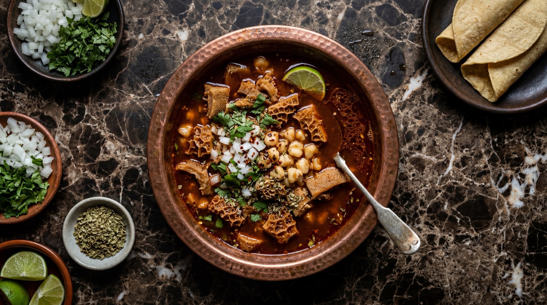 Menudo mexicano rojo con panza de res, maiz pozolero y chile guajillo en cazuela de barro sobre mesa de marmol negro