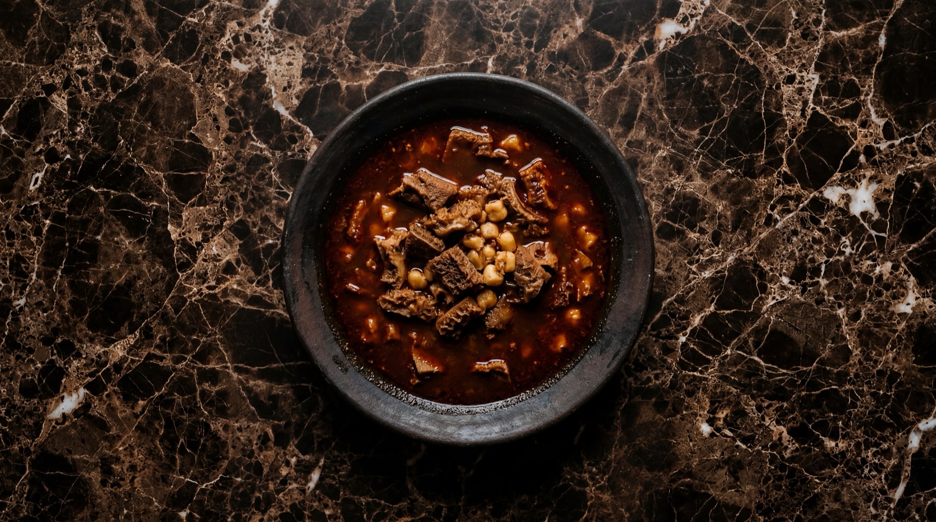 Menudo mexicano rojo con pancita de res y maiz pozolero en tazon de barro sobre mesa de marmol negro