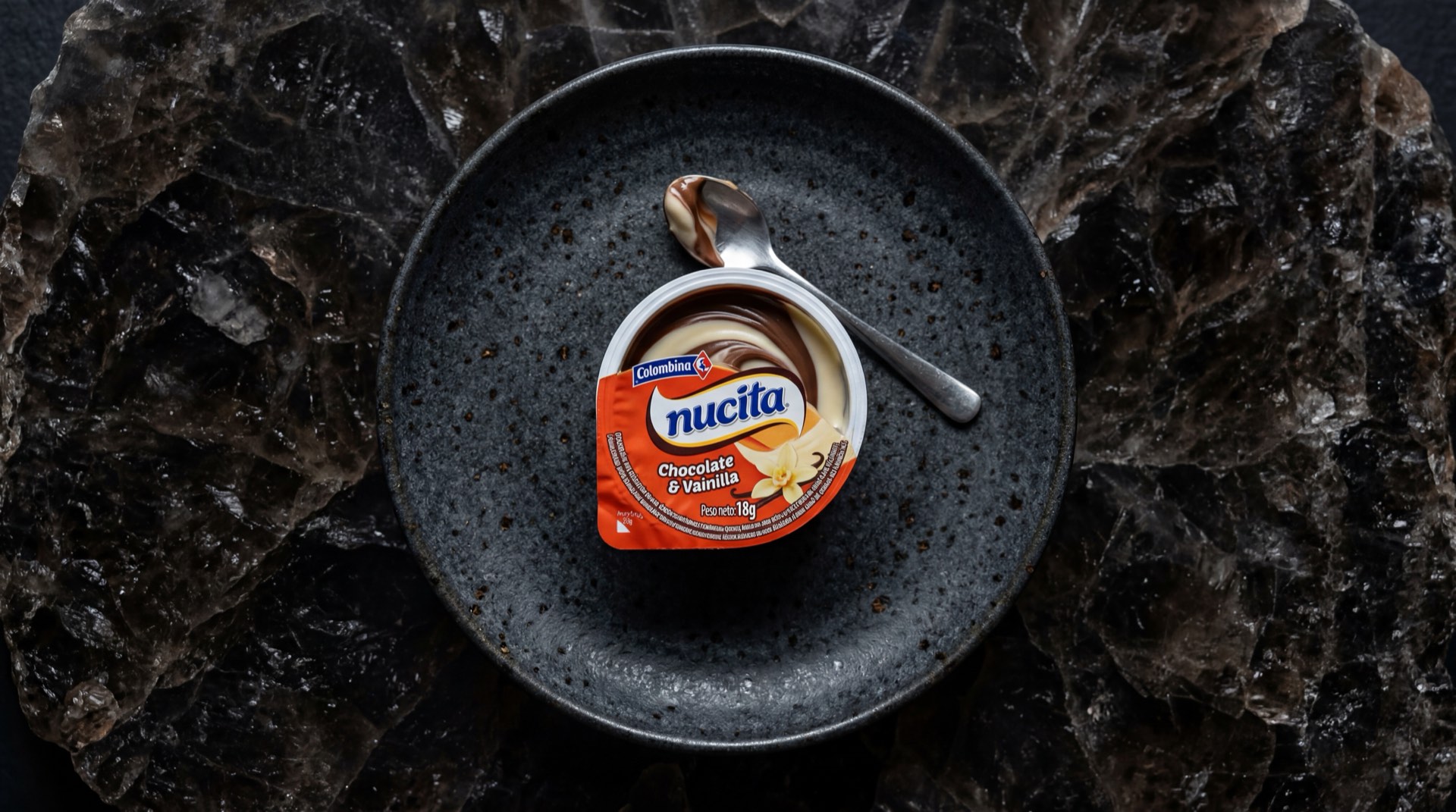 Nucita de Colombina, vasito de crema bicolor de chocolate y vainilla con cucharita sobre mesa de marmol oscuro