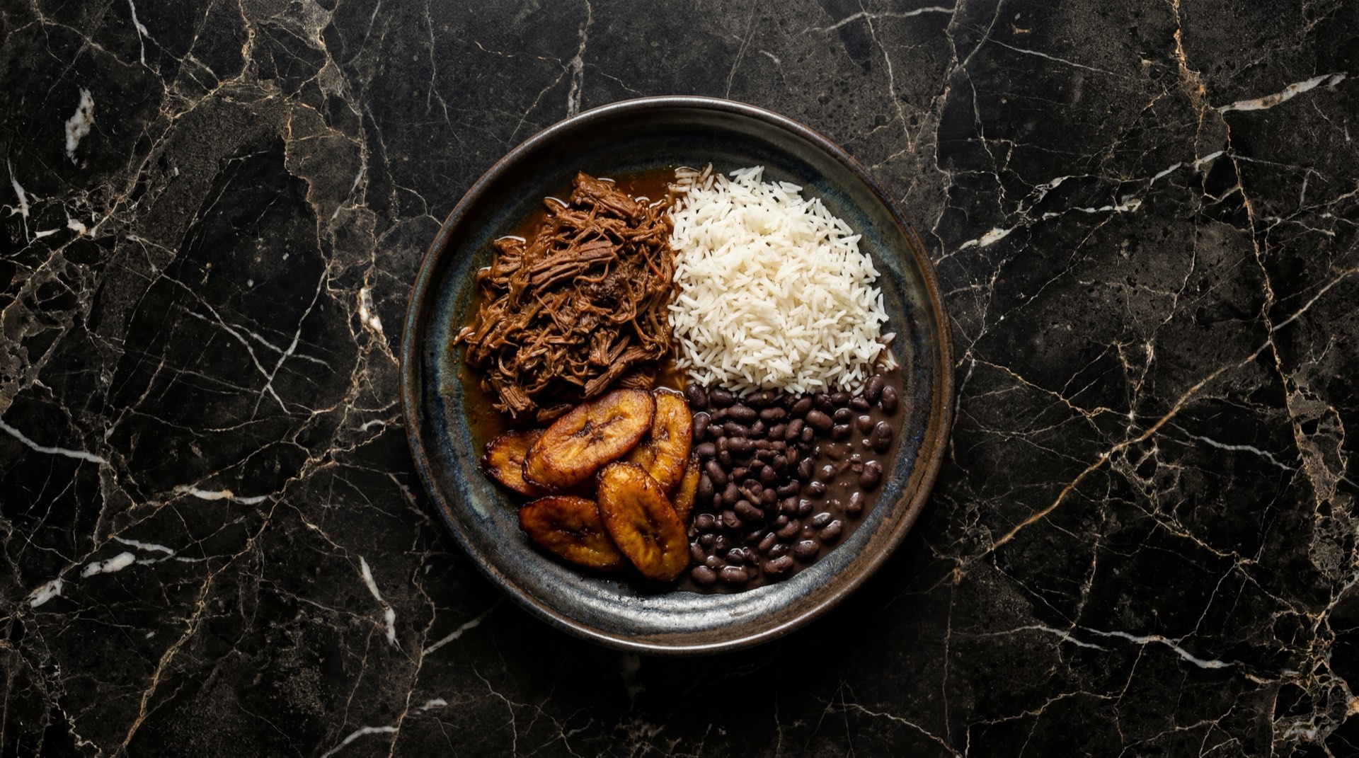 Pabellon criollo venezolano con carne mechada de res desmechada, caraotas negras guisadas, arroz blanco de grano largo y tajadas de platano maduro frito en plato de ceramica oscura sobre mesa de marmol negro con vetas doradas