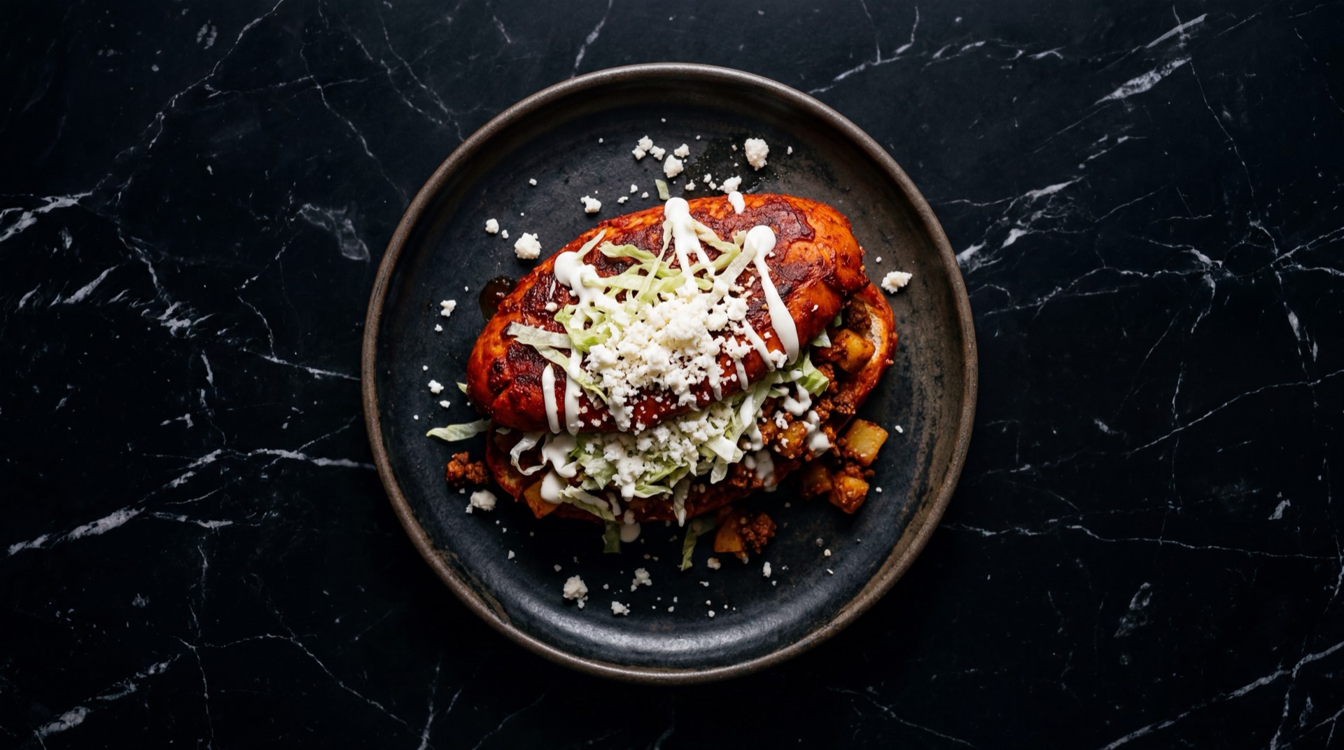 Pambazo mexicano rojo banado en salsa de chile guajillo relleno de papa con chorizo sobre plato de ceramica negra mate en mesa de marmol negro con vetas blancas