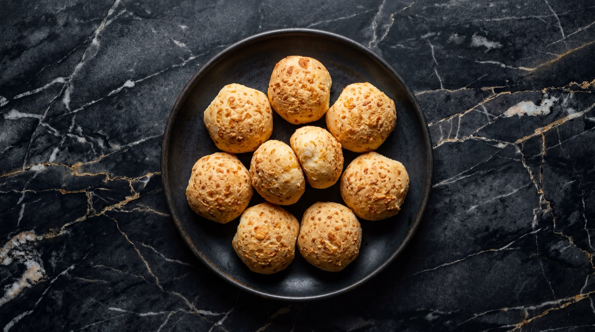 Pandebono valluno recien horneado dorado crujiente en plato de ceramica negro sobre mesa de marmol negro con vetas doradas