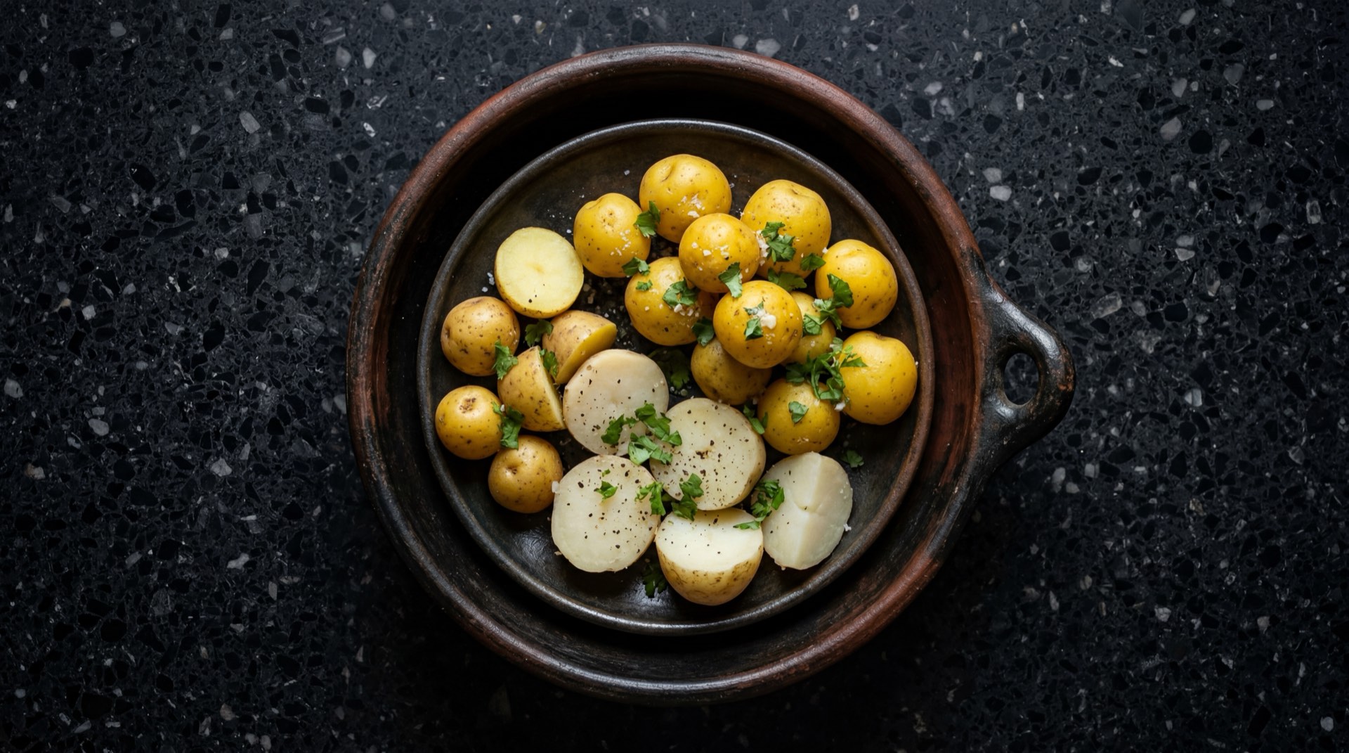 Papas colombianas cocidas enteras con piel dorada en plato de piedra volcanica sobre mesa de granito negro con vetas doradas