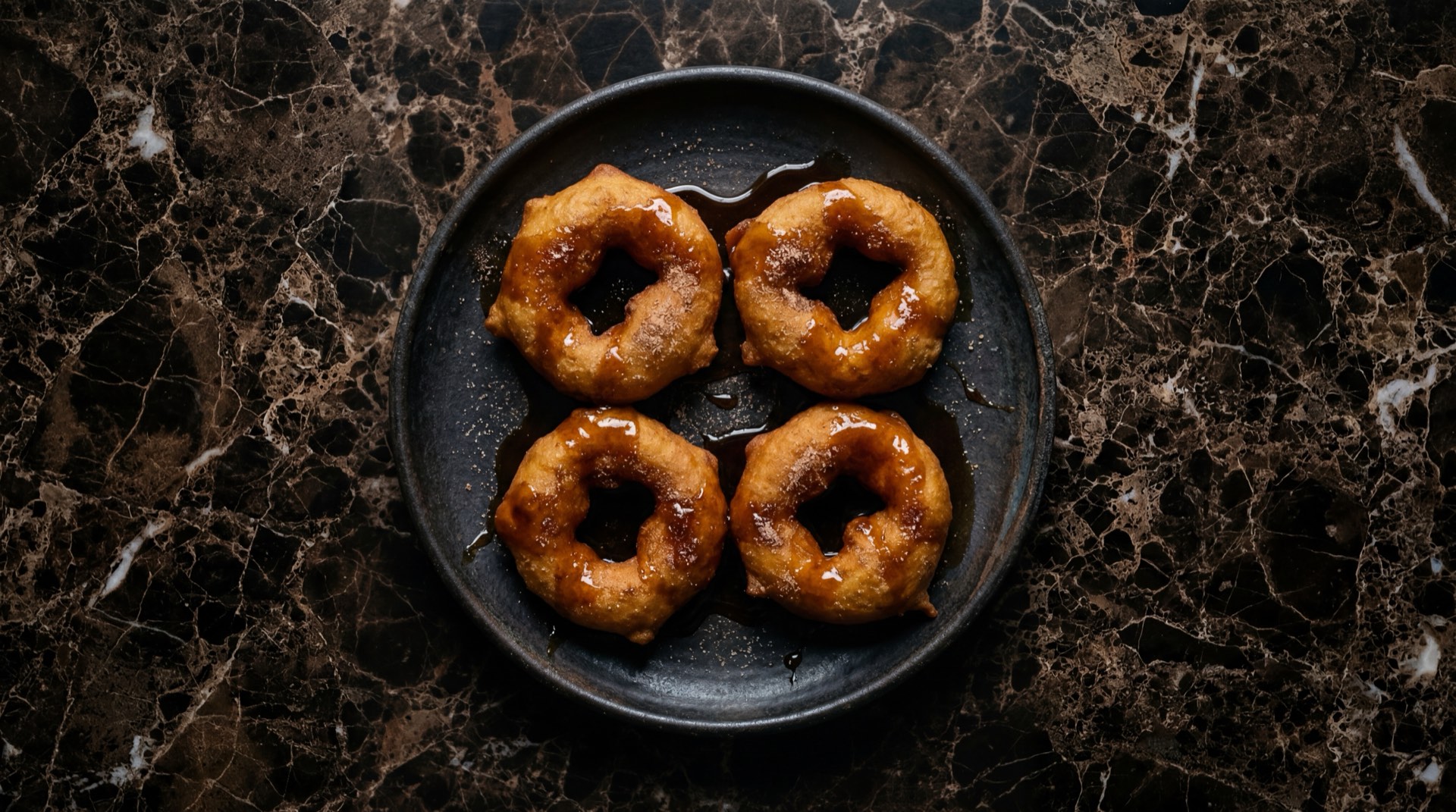 Picarones peruanos dorados en forma de dona banados con miel de chancaca oscura sobre plato de basalto volcanico en mesa de jade verde oscuro