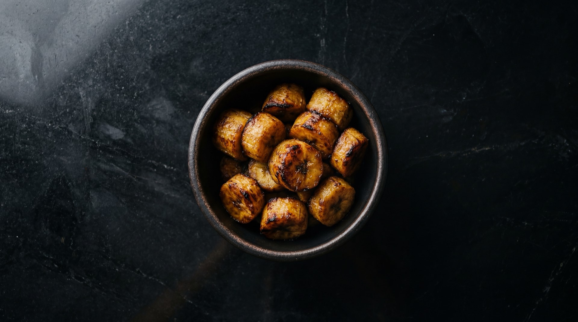 Platano verde harton colombiano cocido en rodajas sobre plato de ceramica mate negro, version saludable sin aceite