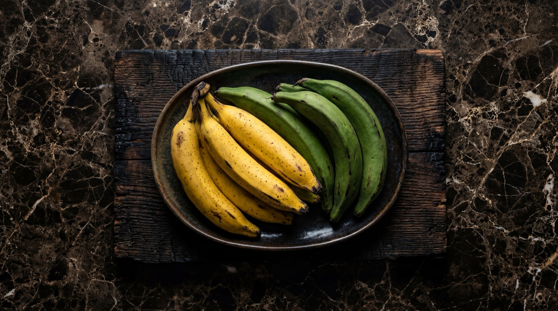 Platano harton colombiano verde y maduro sobre tabla de madera de acacia ahumada en mesa de marmol negro con vetas doradas
