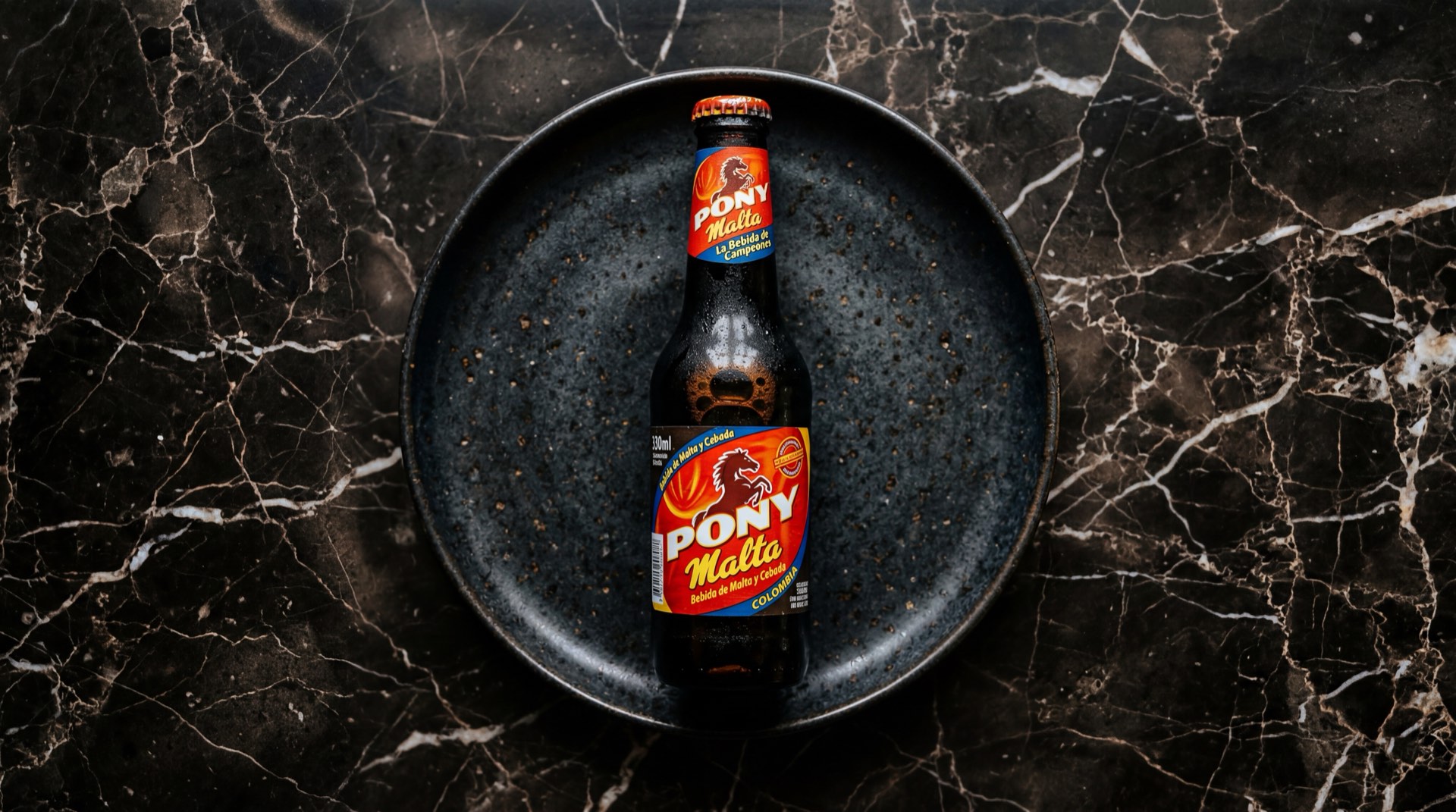 Pony Malta botella de vidrio oscura con liquido marron oscuro espumoso sobre mesa de piedra volcanica negra