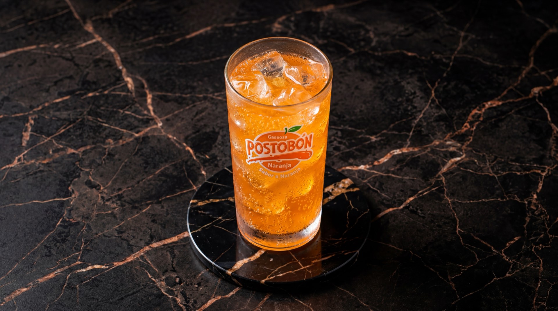 Postobon Naranja botella de vidrio con liquido naranja brillante gasificado sobre mesa de marmol negro con vetas doradas