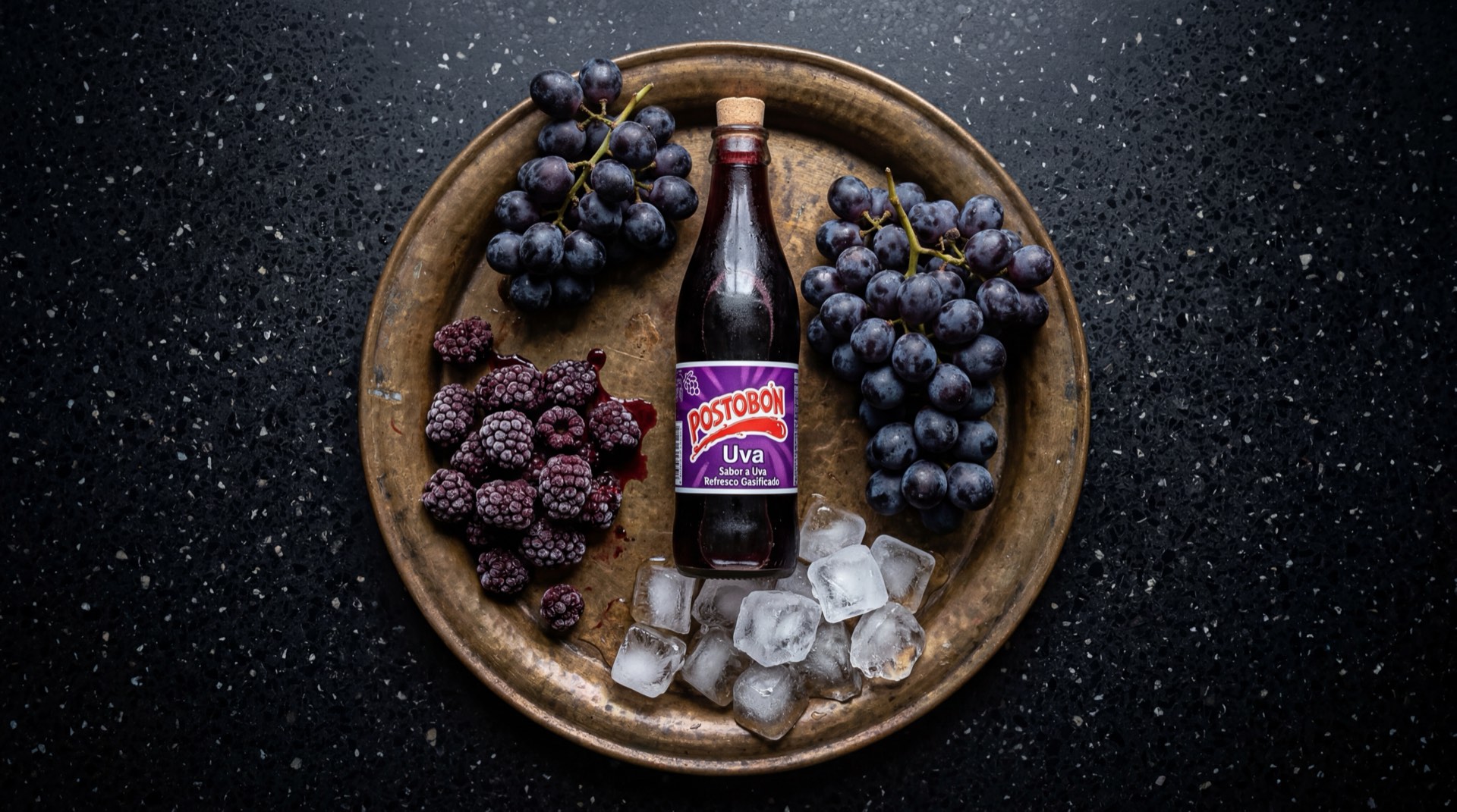 Postobon Uva servida en vaso de vidrio con hielo mostrando el liquido morado intenso gasificado, con azucar refinada blanca y uvas moradas frescas al lado