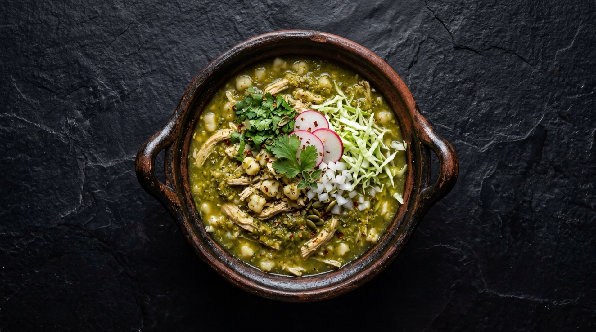 Pozole verde mexicano de pollo con maiz cacahuazintle en plato hondo de ceramica oscura sobre mesa de granito negro con destellos dorados