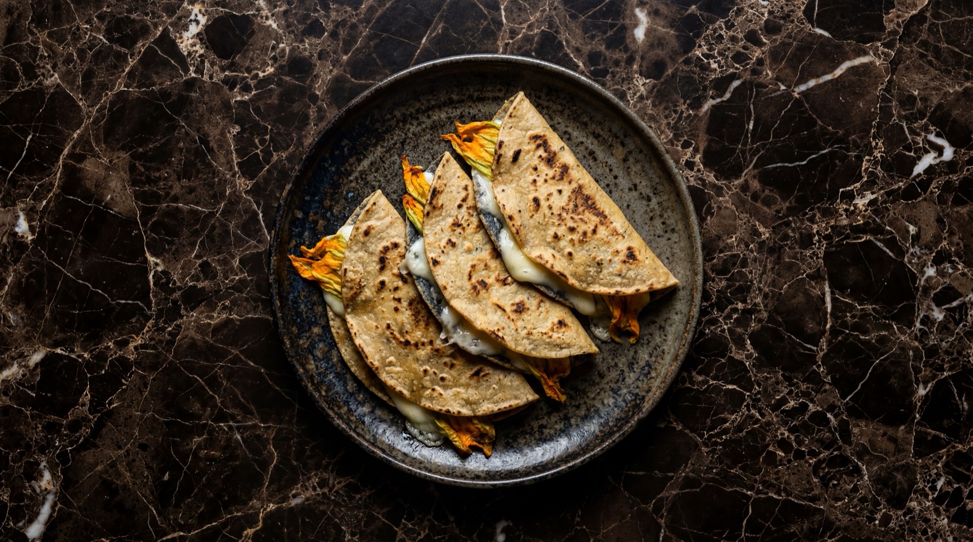 Quesadillas mexicanas de queso Oaxaca derretido en tortilla de maiz sobre plancha de hierro fundido en mesa de marmol negro con vetas doradas