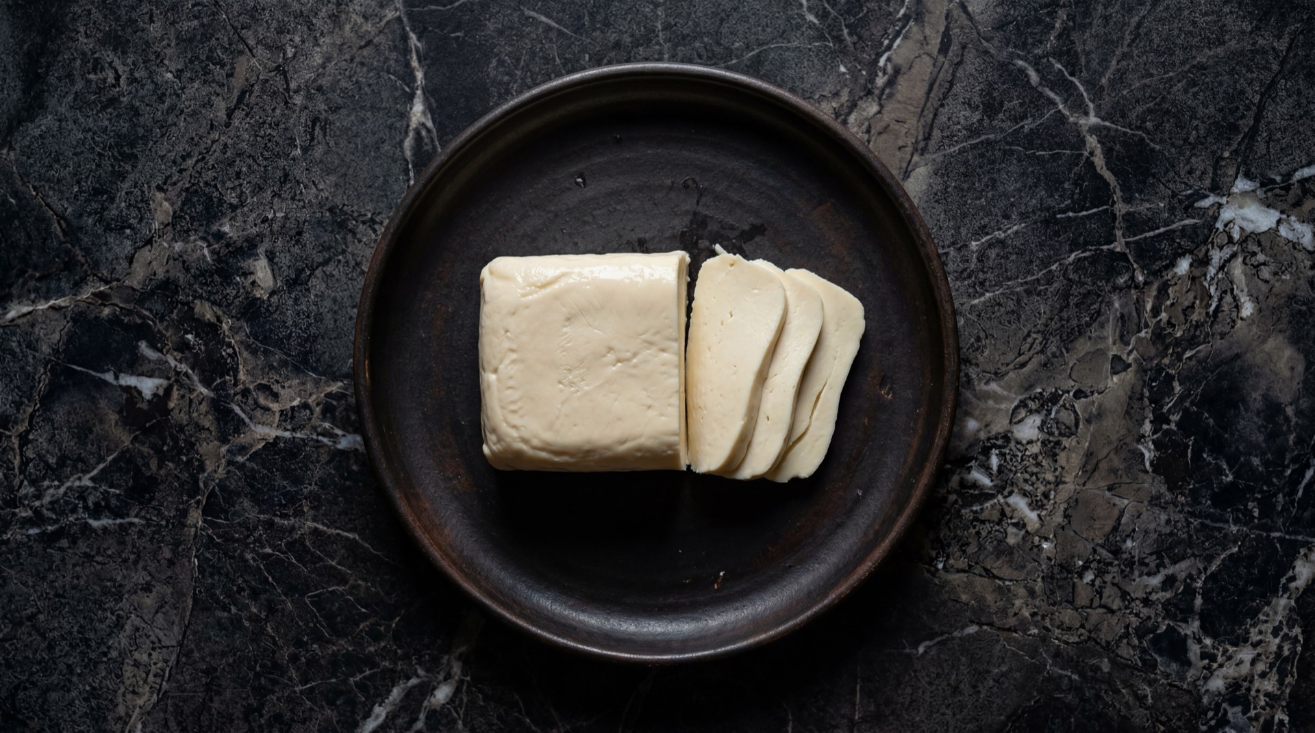 Queso doble crema colombiano semi-blando derritiendose sobre tabla de madera de acacia en mesa de granito negro con destellos dorados