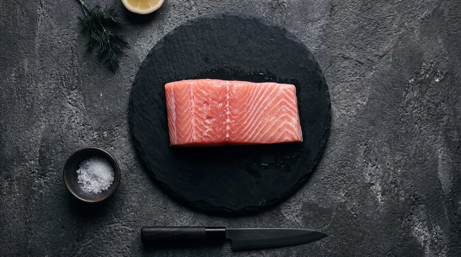 Filete de salmon fresco al horno con limon verde y eneldo sobre plato de ceramica negra mate en mesa de marmol negro con vetas doradas