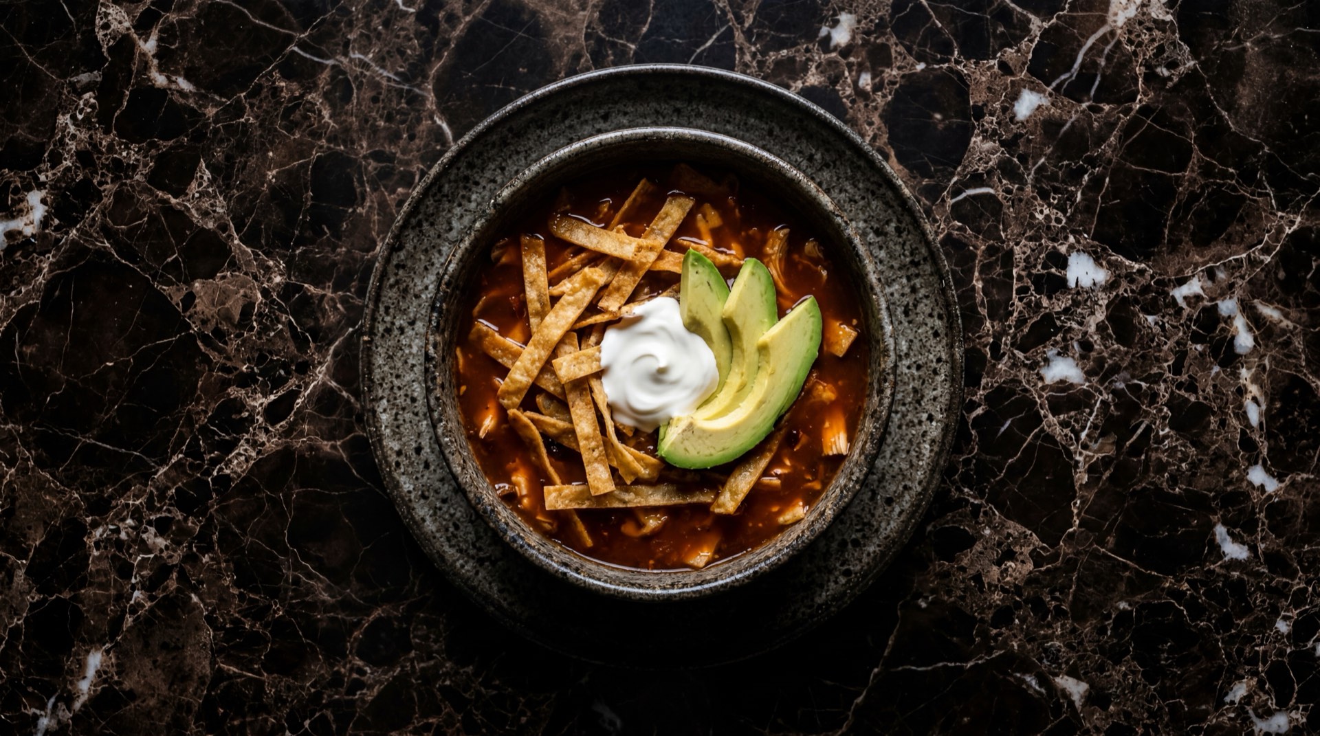 Sopa azteca mexicana con tiras de tortilla fritas crujientes y aguacate maduro en tazon hondo de barro negro sobre mesa de piedra volcanica oscura