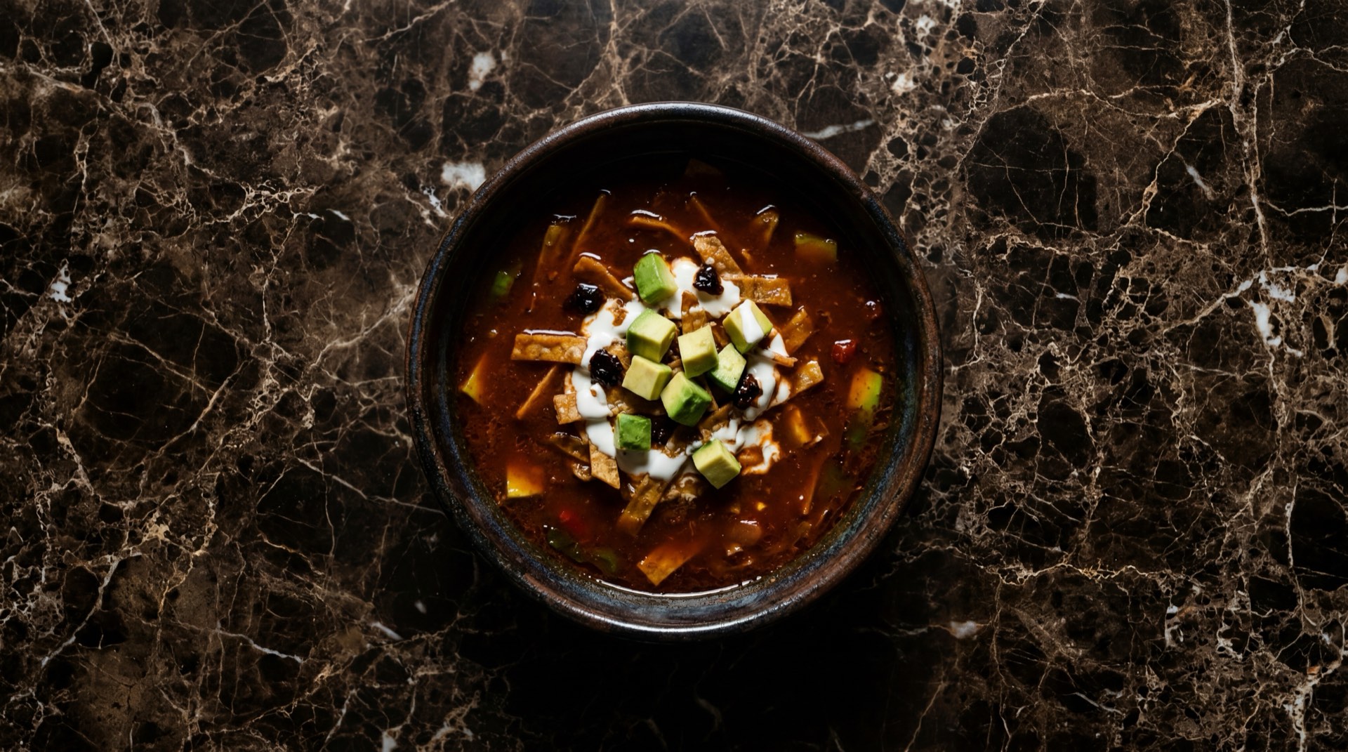 Sopa de tortilla mexicana con tiras de tortilla de maiz frita, aguacate Hass, crema acida y chile pasilla en plato de barro sobre mesa de granito oscuro