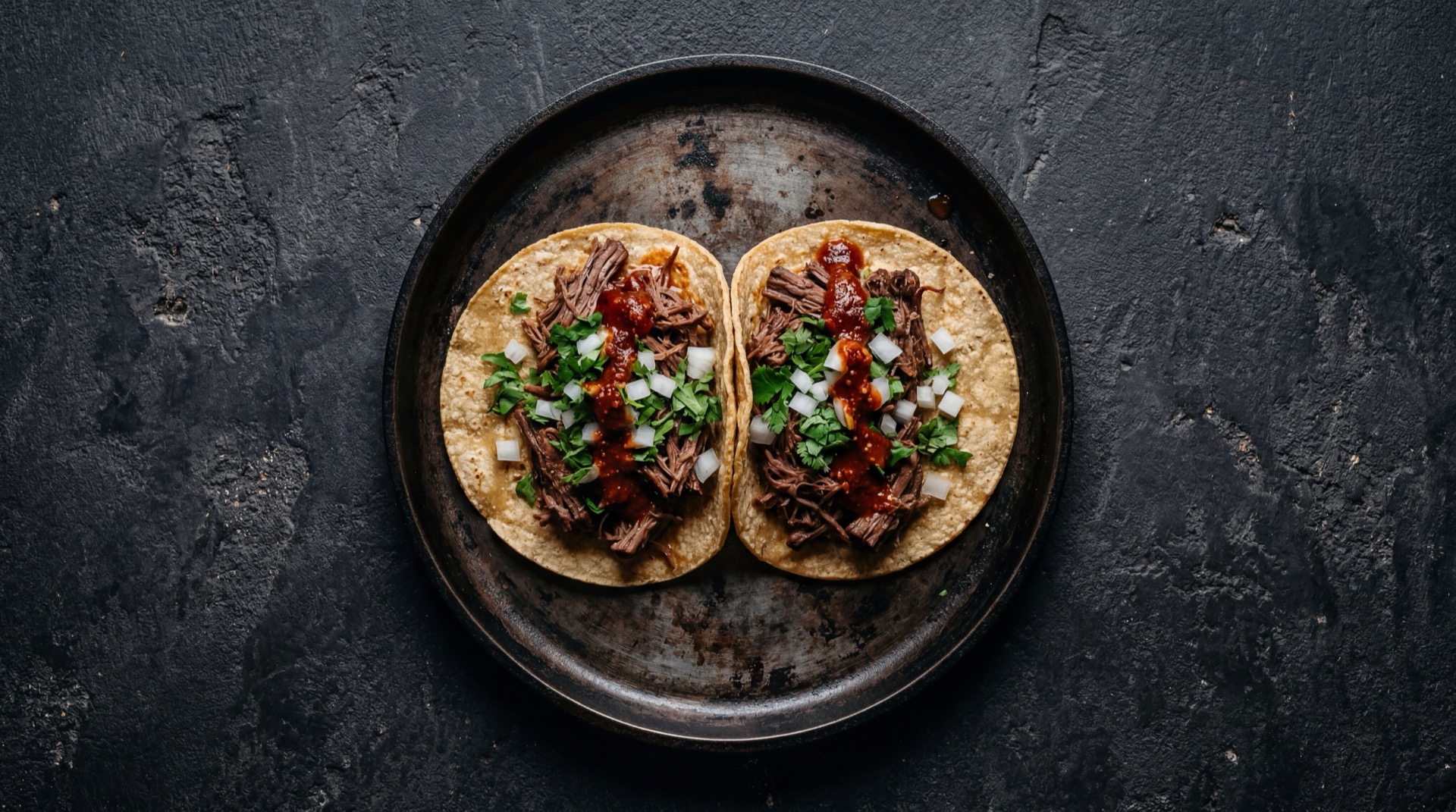 Tacos de barbacoa de borrego desmenuzada con cilantro fresco picado y cebolla blanca picada sobre tortillas de maiz frescas en plato de hierro forjado sobre mesa de granito negro con destellos dorados