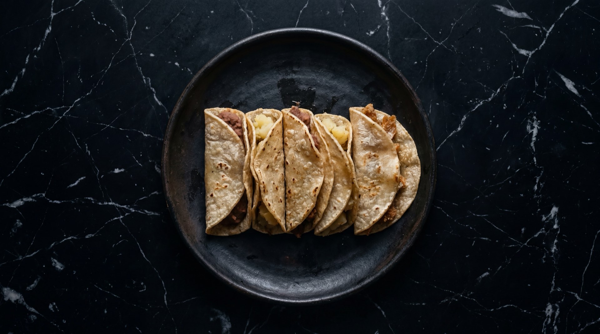 Tacos de canasta mexicanos sudados en canasta de mimbre forrada con papel encerado empapado en aceite, con rellenos de frijol refrito, chicharron prensado en salsa verde y papa machacada con chorizo sobre tabla de madera de nogal oscuro