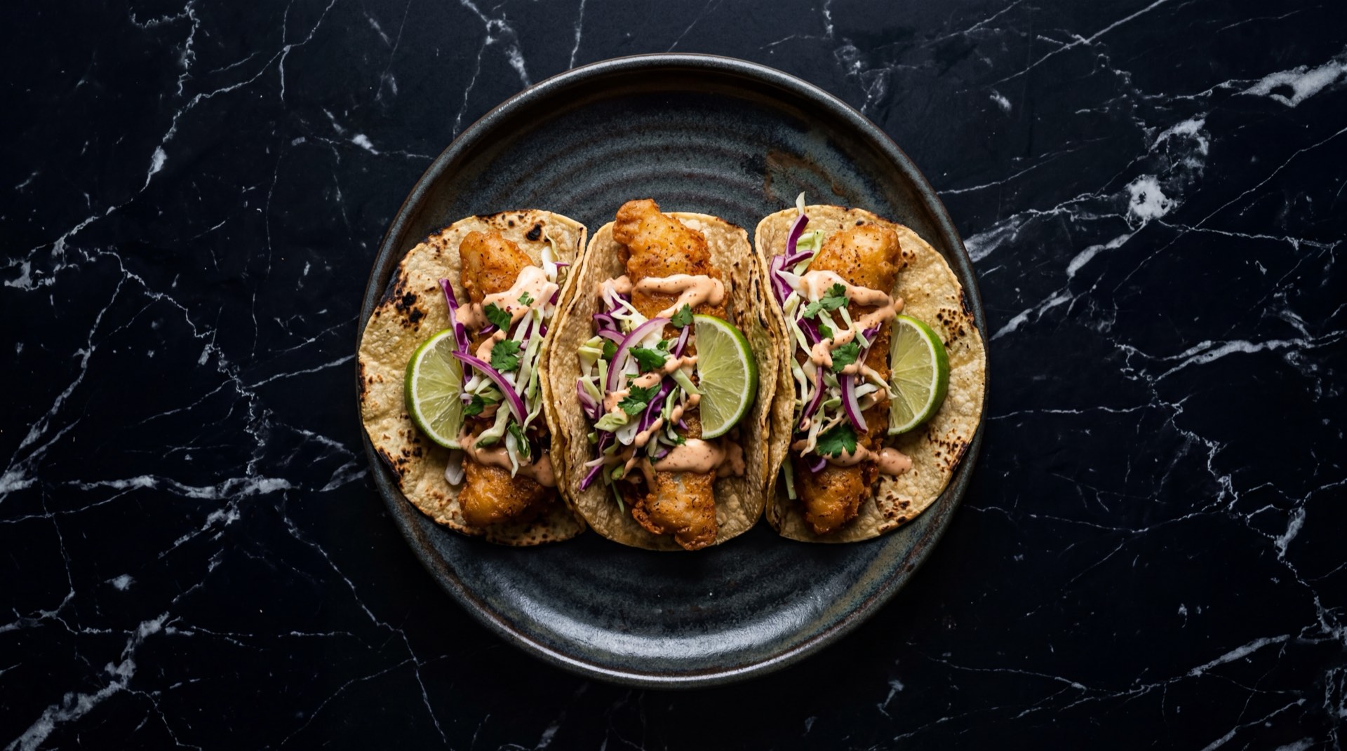 Tacos de pescado estilo Baja California con filete de pescado blanco capeado en masa de cerveza, repollo morado rallado, crema de chipotle adobado, pico de gallo fresco y limon verde sobre plancha de hierro fundido