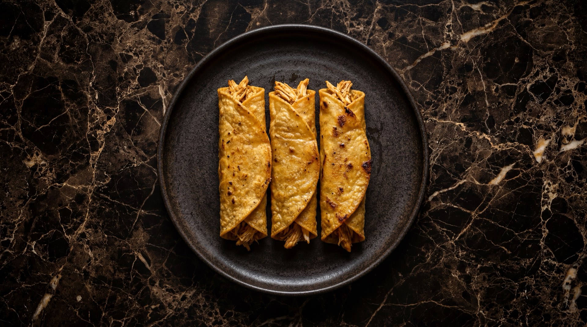 Tacos dorados mexicanos crujientes de pollo desmenuzado con lechuga iceberg rallada, crema agria mexicana y salsa verde casera de tomatillo sobre bandeja de acero ennegrecido
